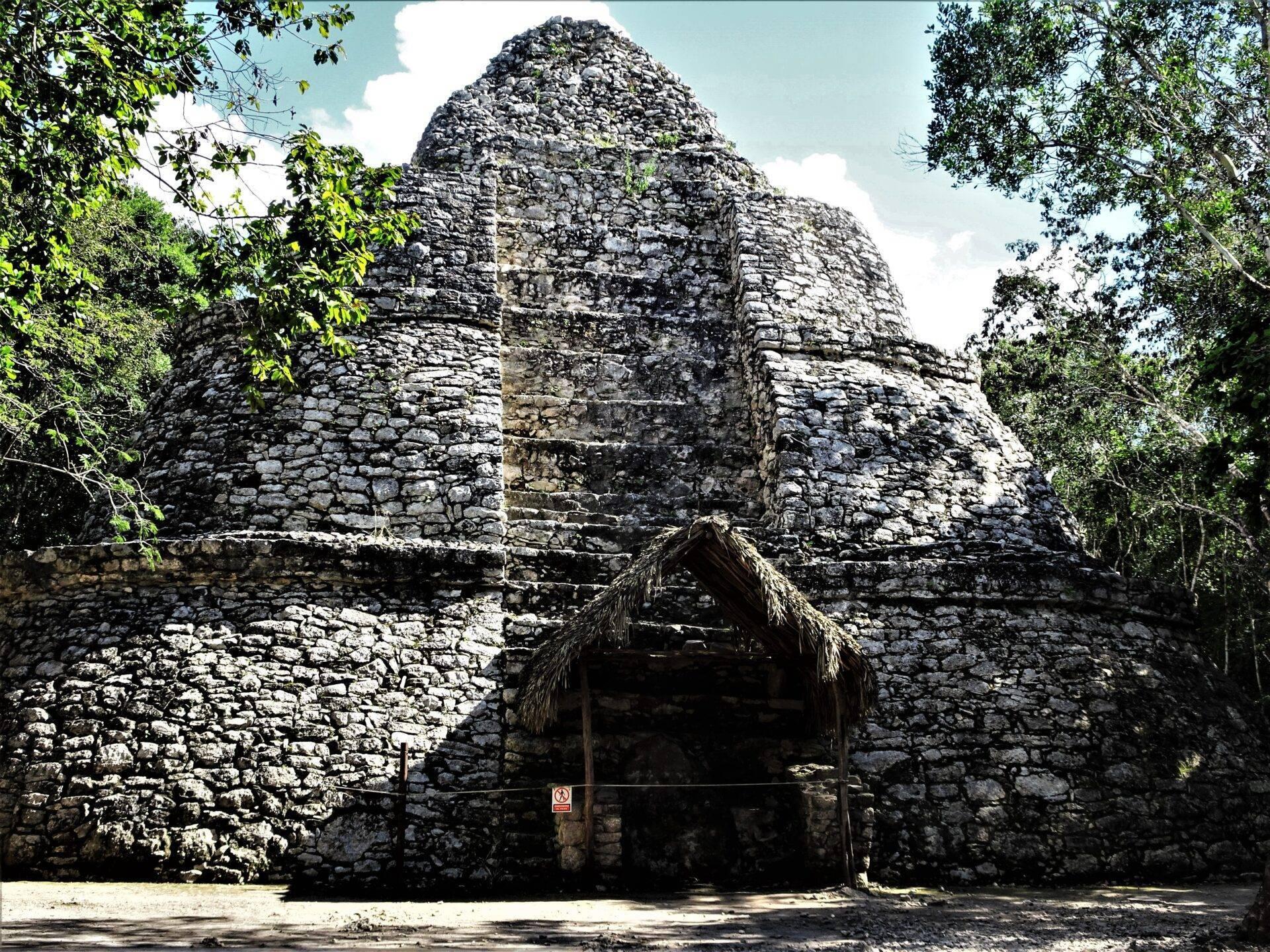 Ruine maya en pierre avec pyramide centrale entourée de végétation tropicale et abri de chaume à l’entrée