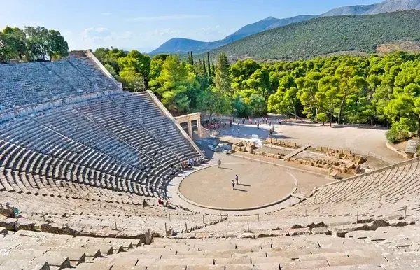Un ancien théâtre antique en pierre avec gradins semi-circulaires, entouré de verdure et montagnes méditerranéennes
