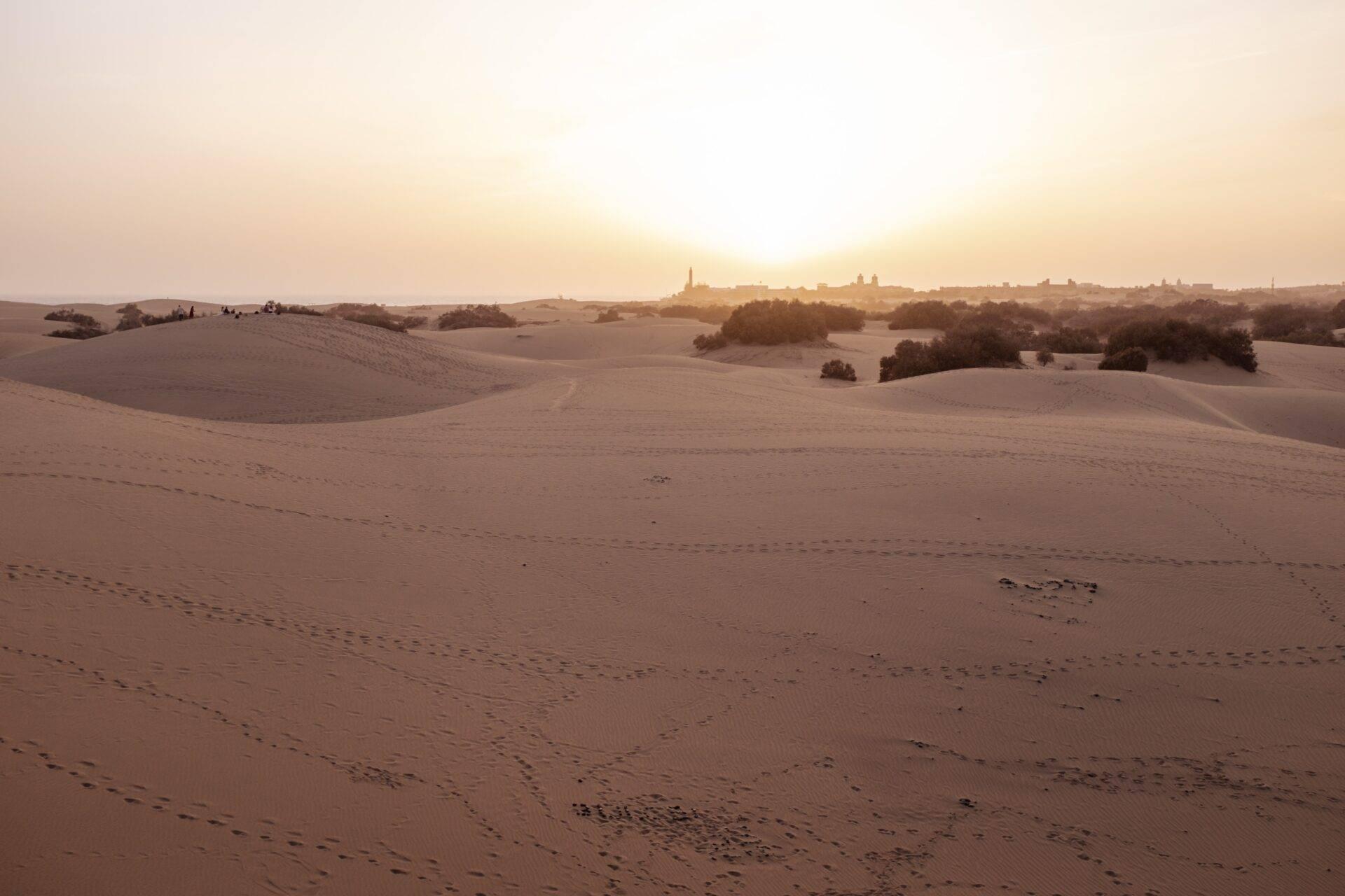 Paysage désertique avec dunes ondulantes et village fortifié illuminé à l’horizon au coucher du soleil