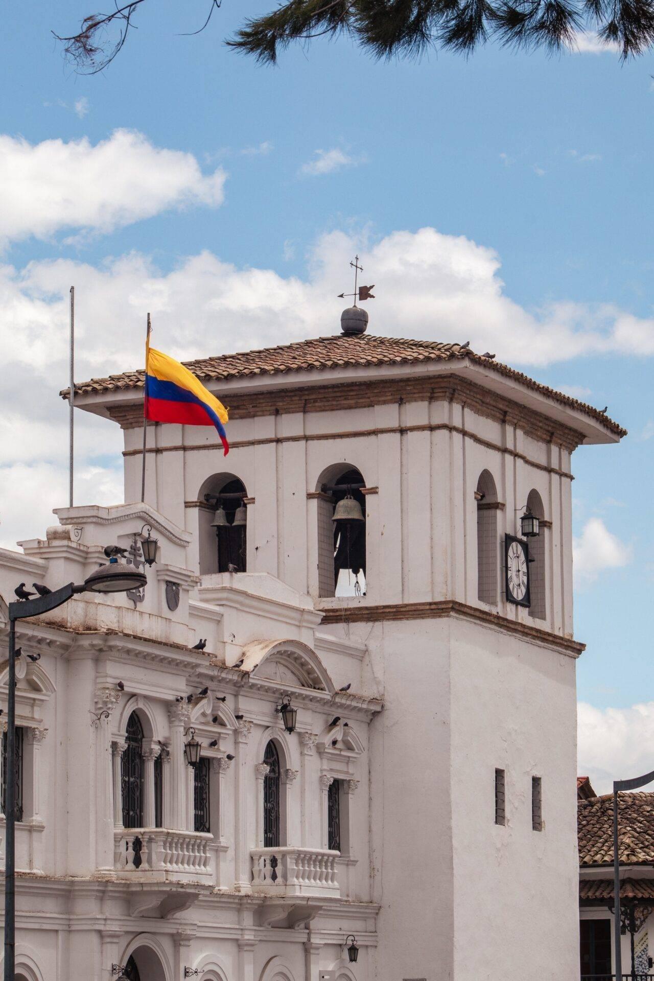 Église coloniale blanche avec clocher et drapeau colombien flottant sous un ciel nuageux