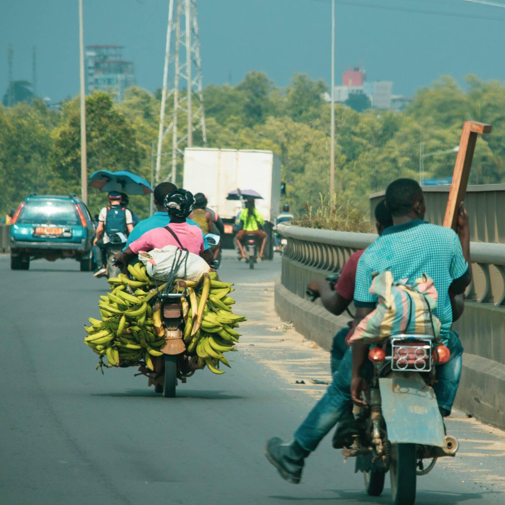 Deux motocyclistes transportent des régimes de bananes sur une route de ville camerounaise animée avec circulation