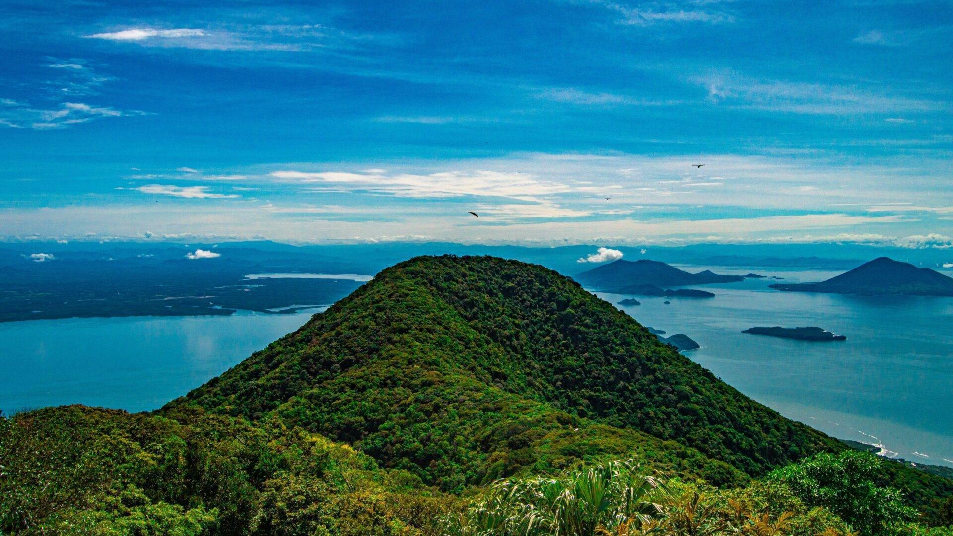 Vue aérienne d’une île densément boisée entourée d’une mer turquoise avec îlots et montagnes en arrière-plan