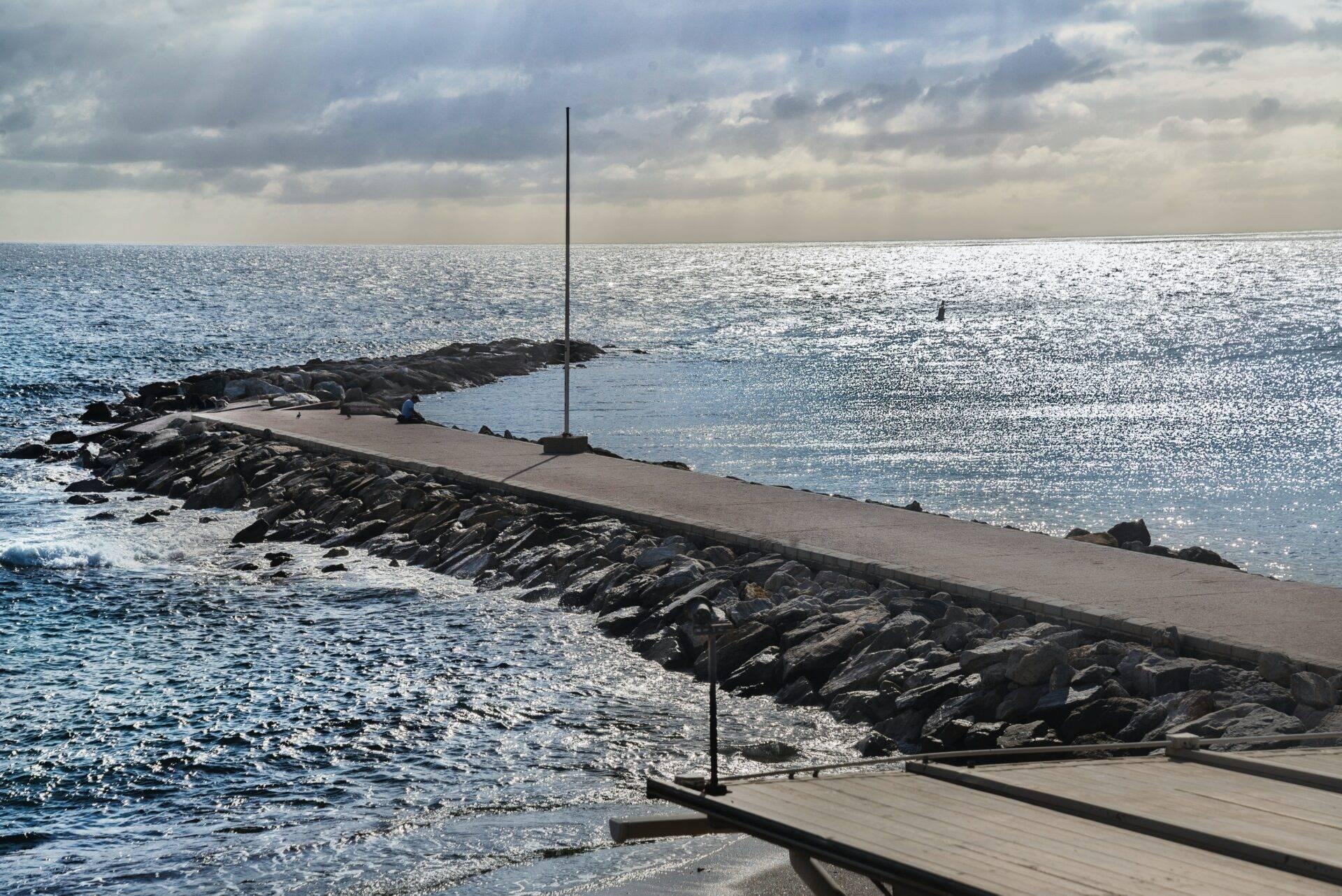 Une jetée en béton avec un mât s’étend vers la mer, bordée de rochers, par temps nuageux