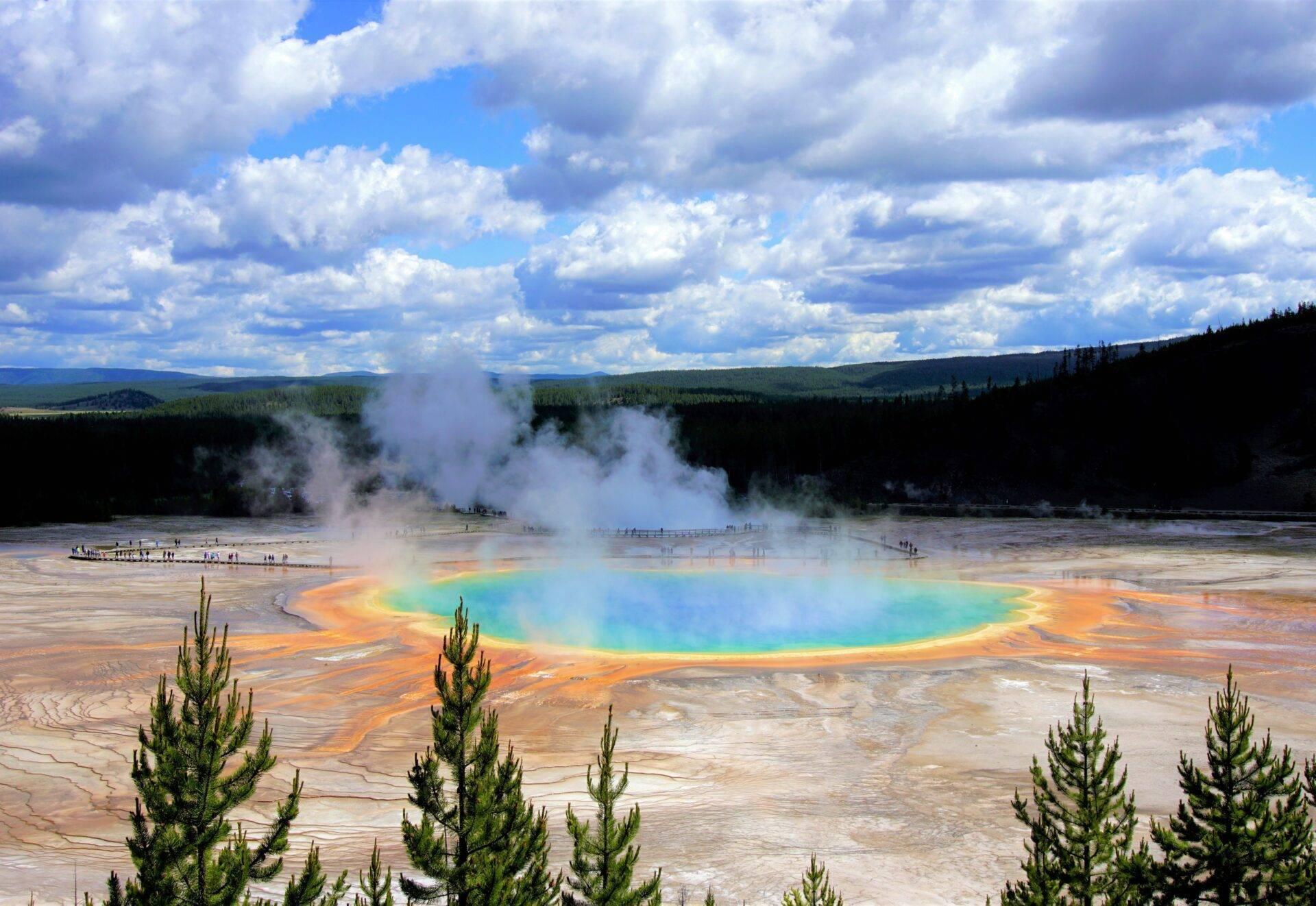 Un geyser géothermique aux eaux turquoise et orange entouré de forêt de conifères sous un ciel nuageux