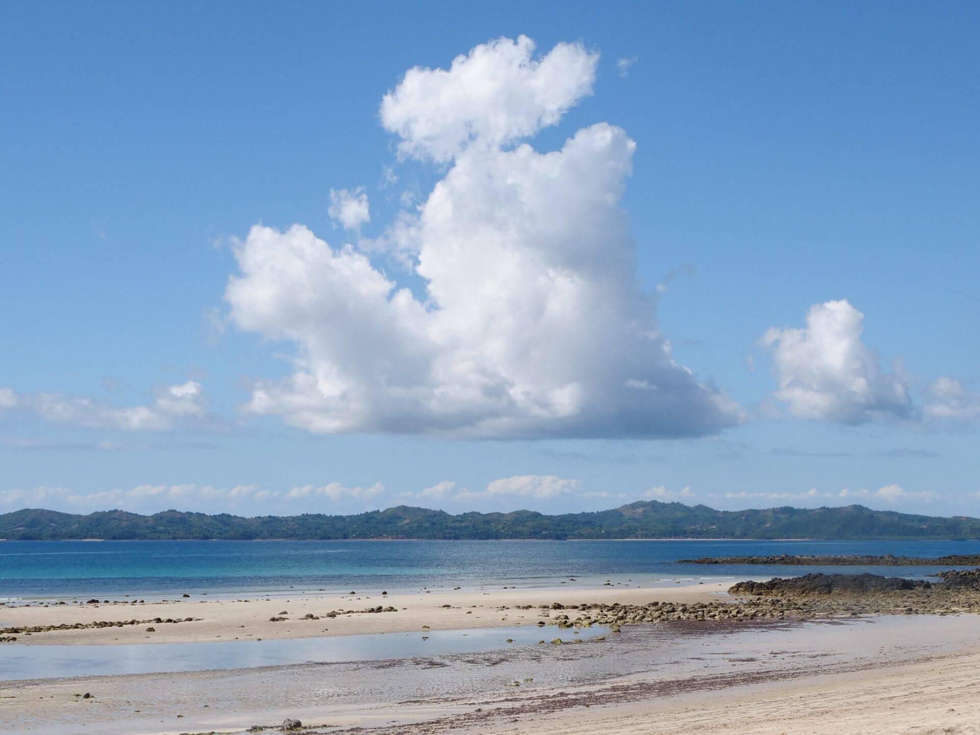 Une plage de sable blanc avec lagon turquoise, montagnes boisées en arrière-plan et ciel bleu nuageux