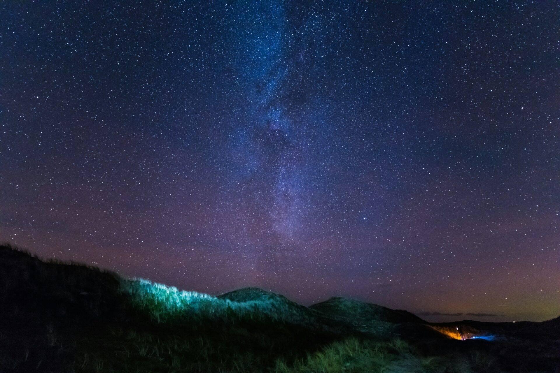 Montagnes boisées sous un ciel étoilé avec la Voie lactée visible et quelques lumières au loin
