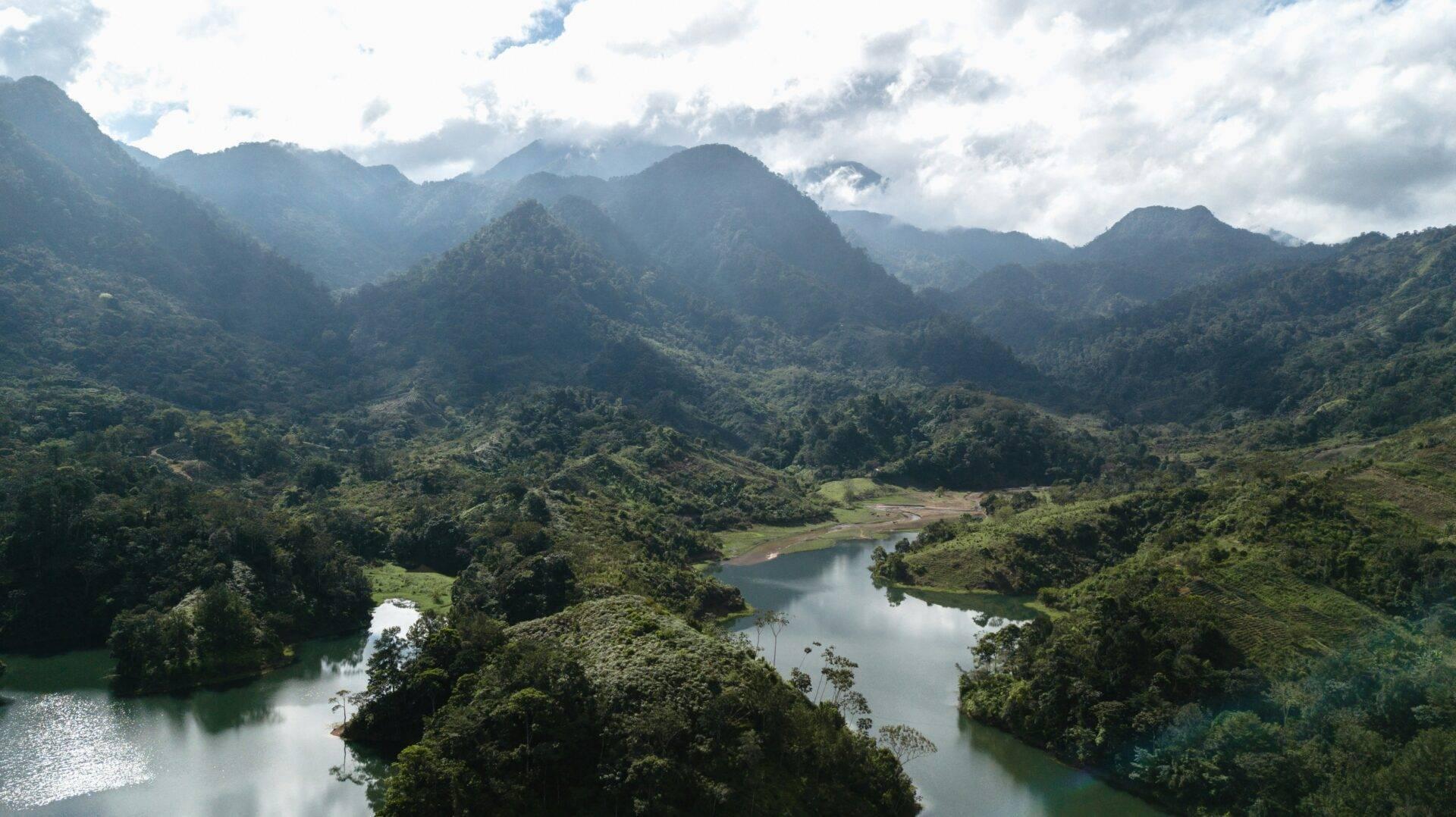 Vallée verdoyante entourée de montagnes boisées avec rivières et lacs en miroir sous un ciel nuageux