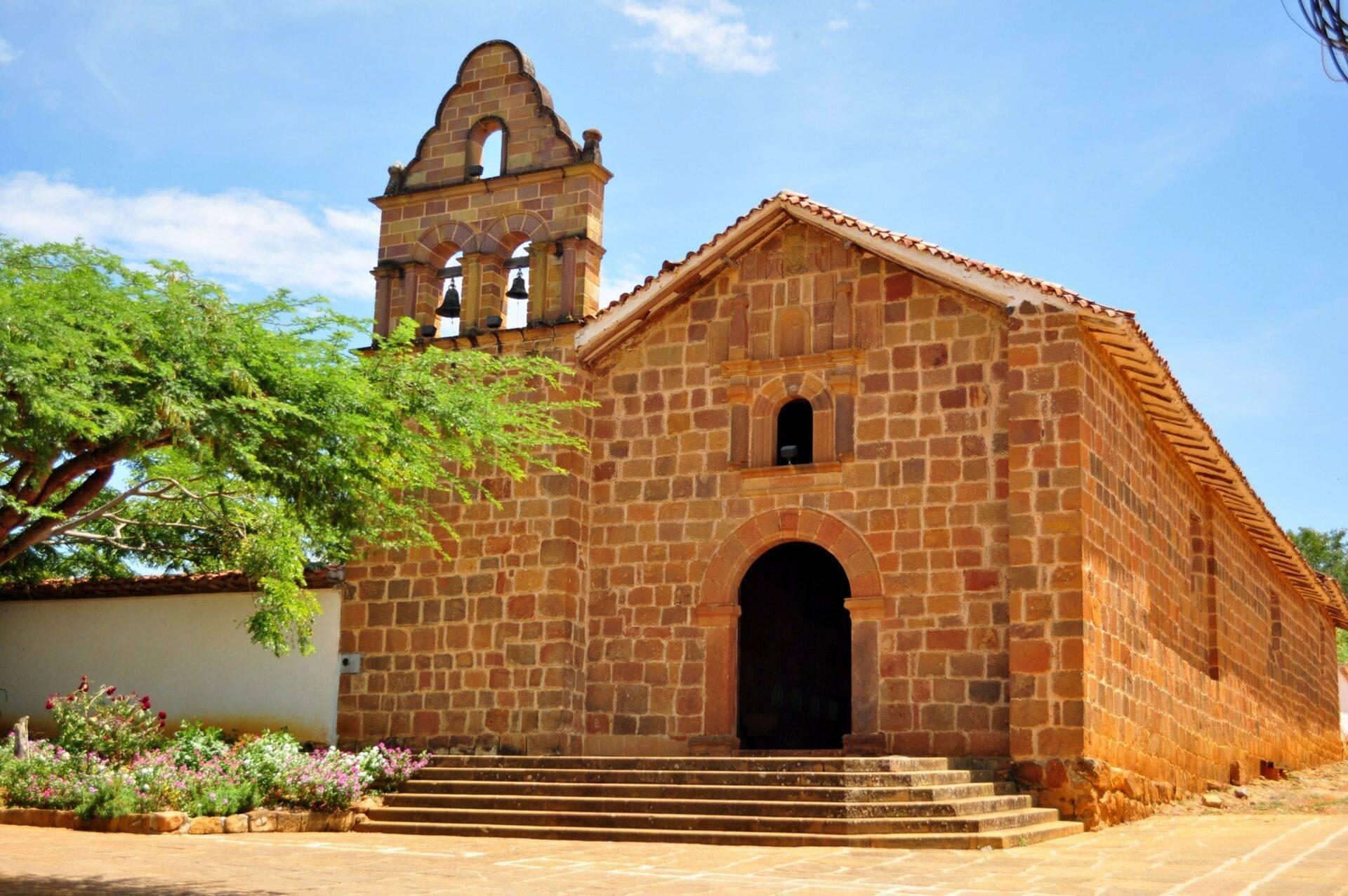 Église coloniale en brique rouge avec clocher à campanile sous ciel bleu, entourée de végétation tropicale