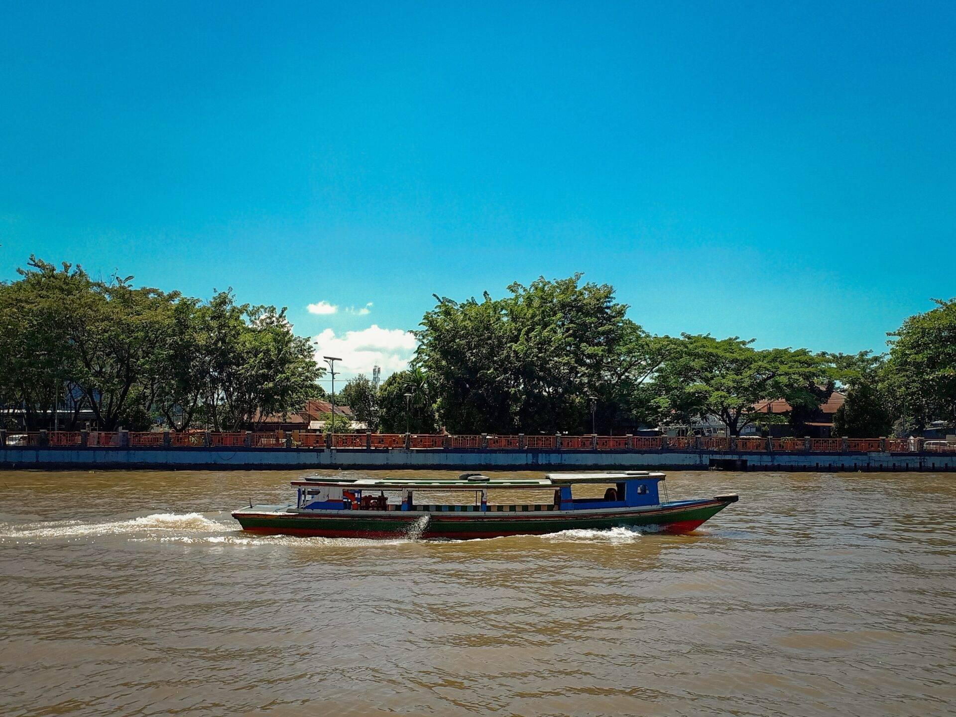 Un bateau de transport fluvial coloré navigue sur un fleuve marron sous un ciel bleu dégagé, avec des arbres et des structures portuaires en arrière-plan