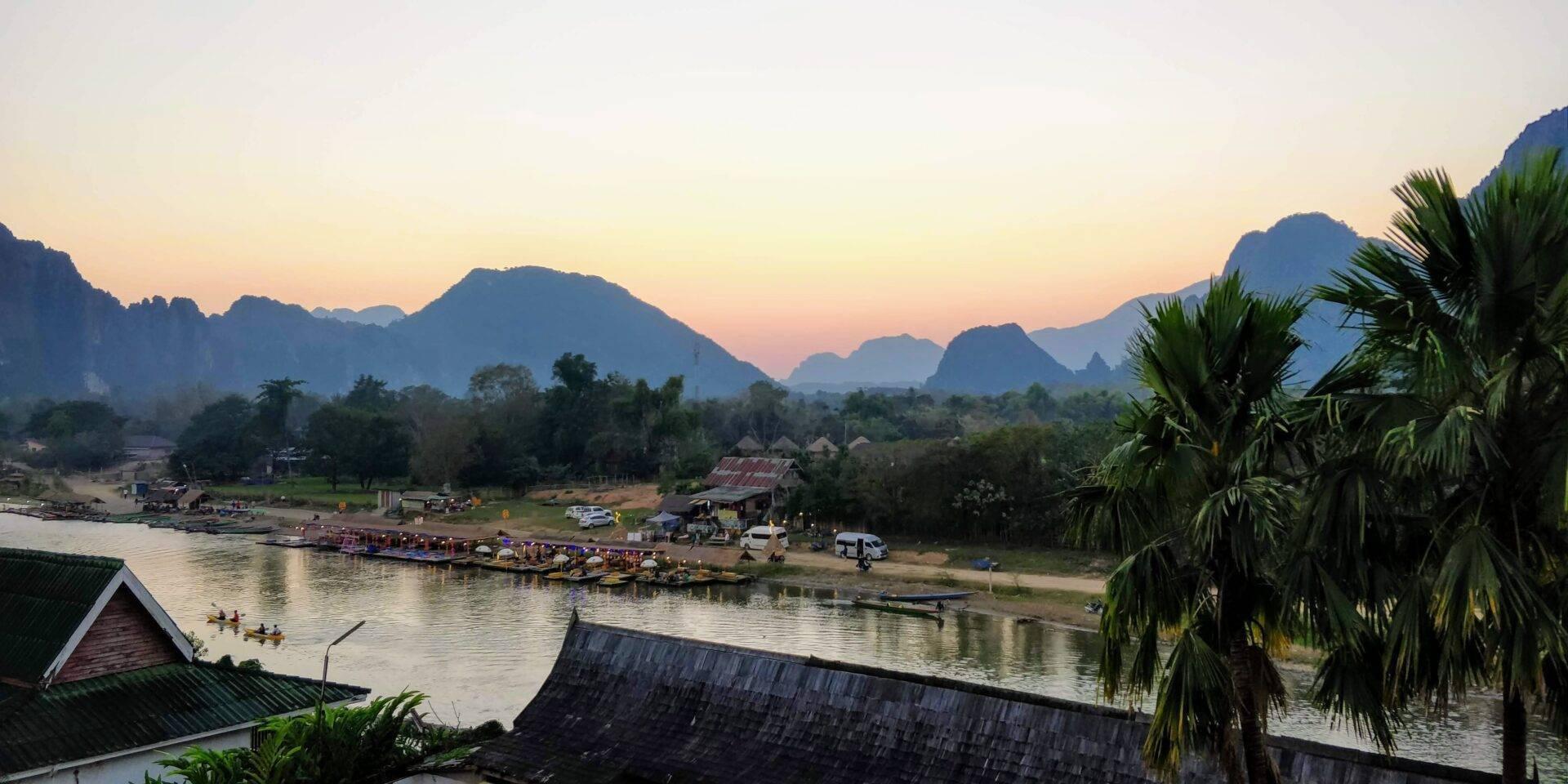 Vue panoramique d’une vallée fluviale en Asie du Sud-Est au crépuscule, avec montagnes calcaires, palmiers et village traditionnel