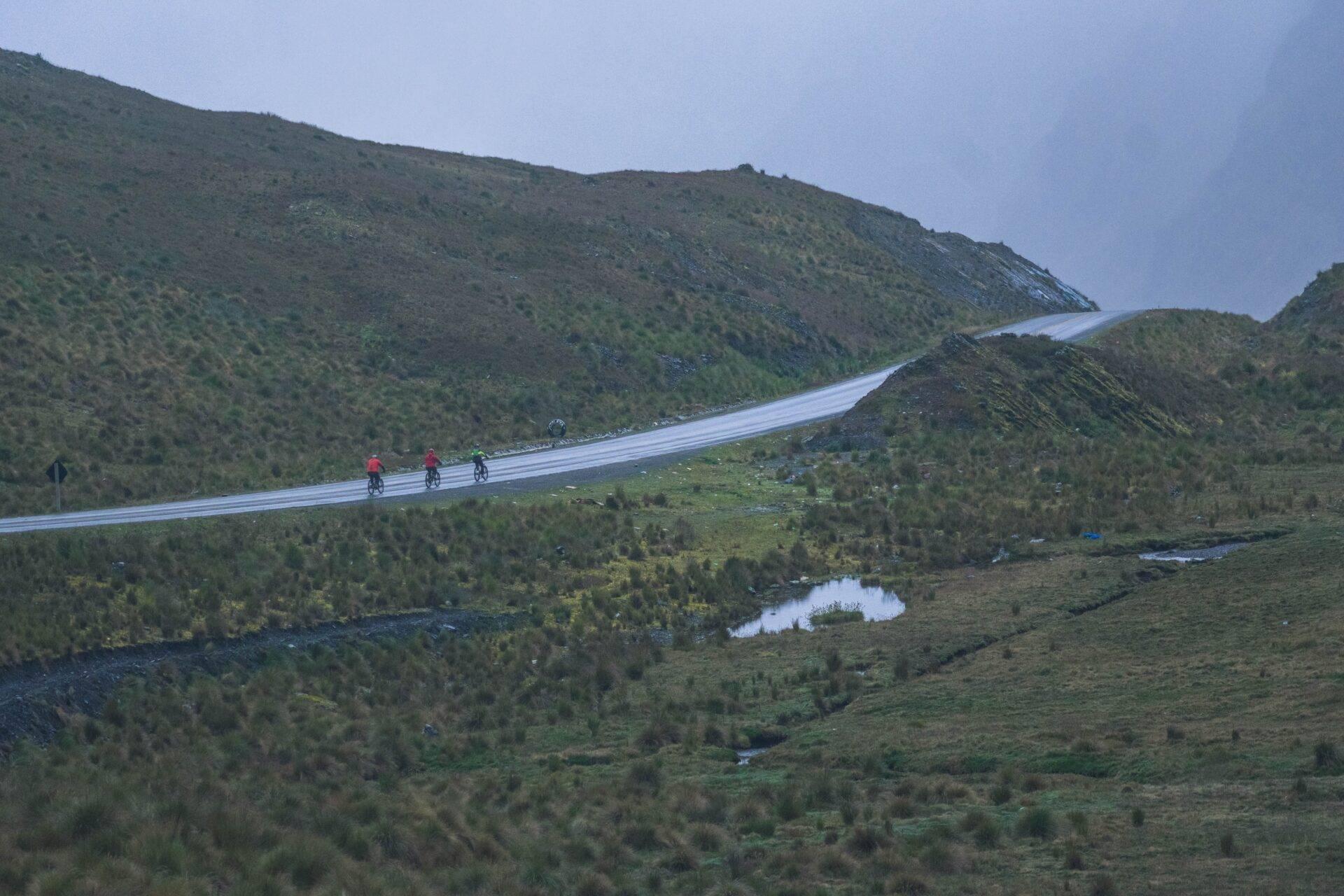 Route asphaltée sinueuse traversant des montagnes boisées avec des cyclistes et un paysage de toundra alpine