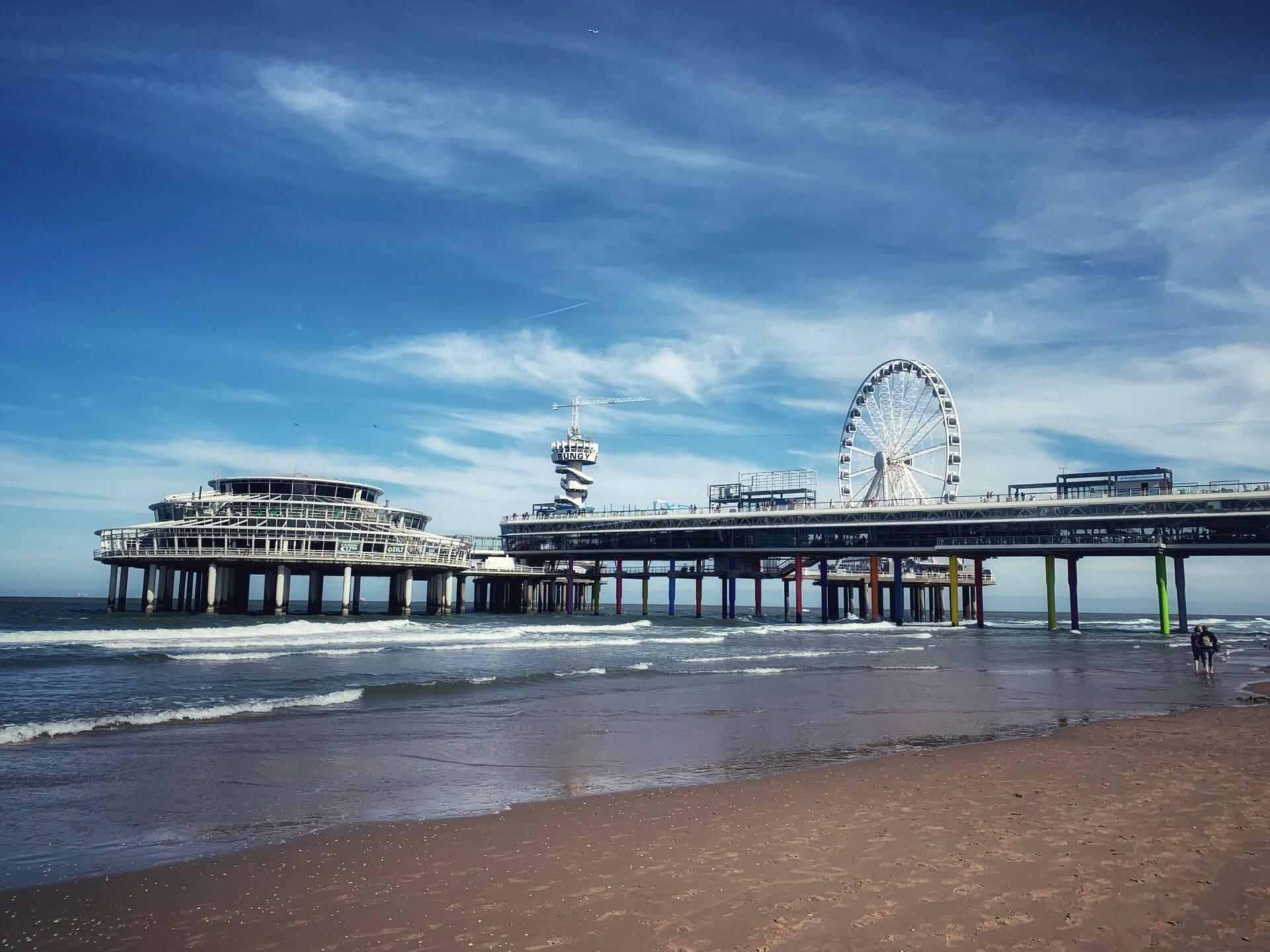 Pier côtier avec grande roue, attractions et plage de sable sous ciel bleu nuageux