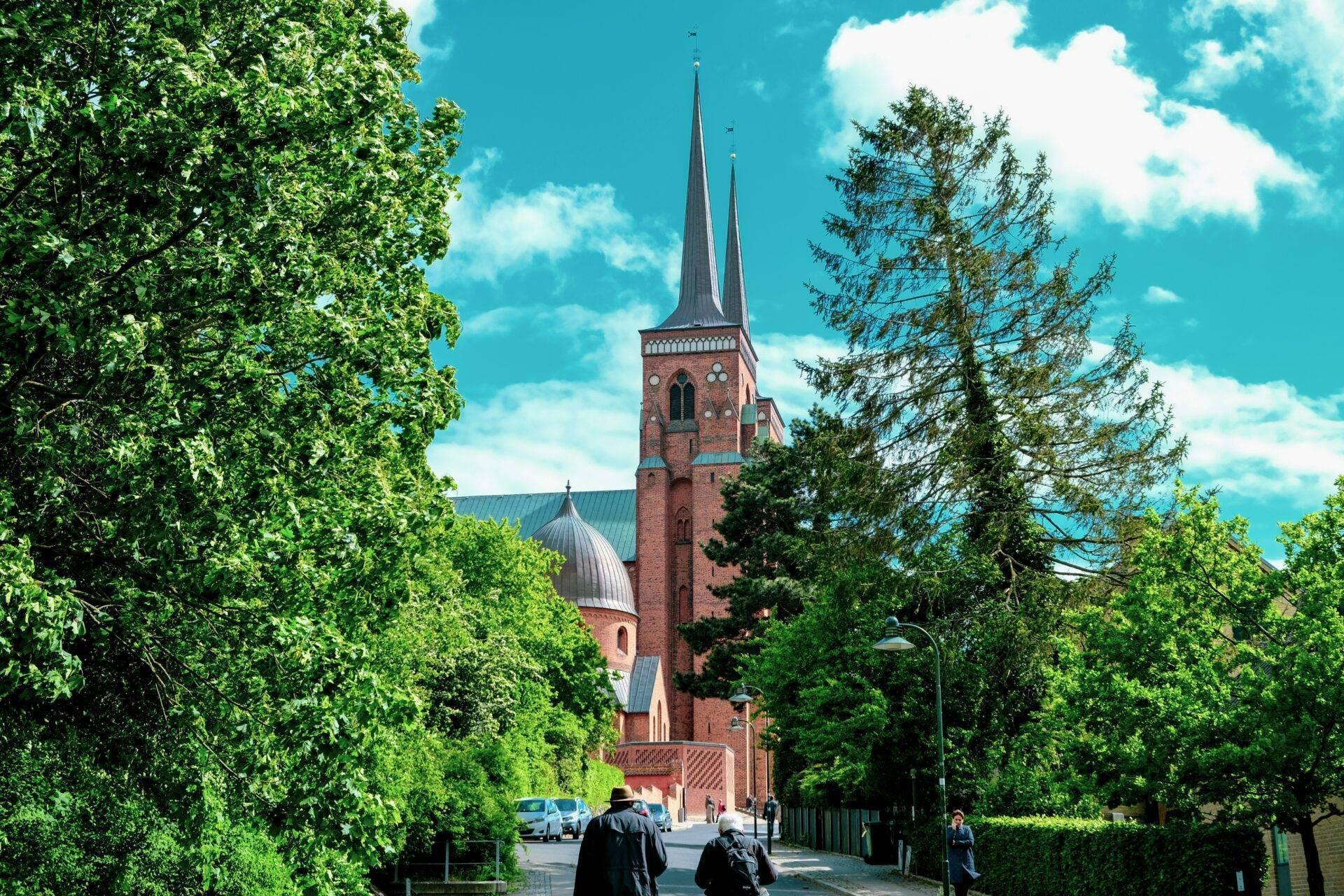 Une église de briques rouges aux deux clochers pointus, entourée de verdure luxuriante et d’une rue paisible