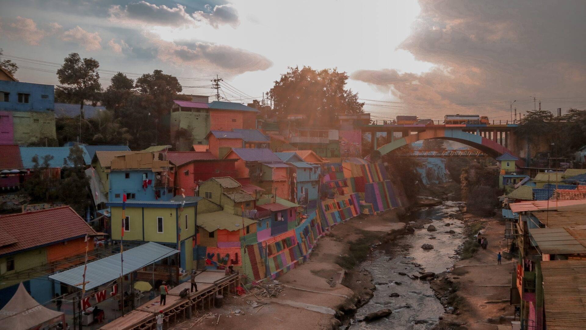 Village coloré au bord d’une rivière avec maisons peintes aux couleurs vives, pont et ciel nuageux