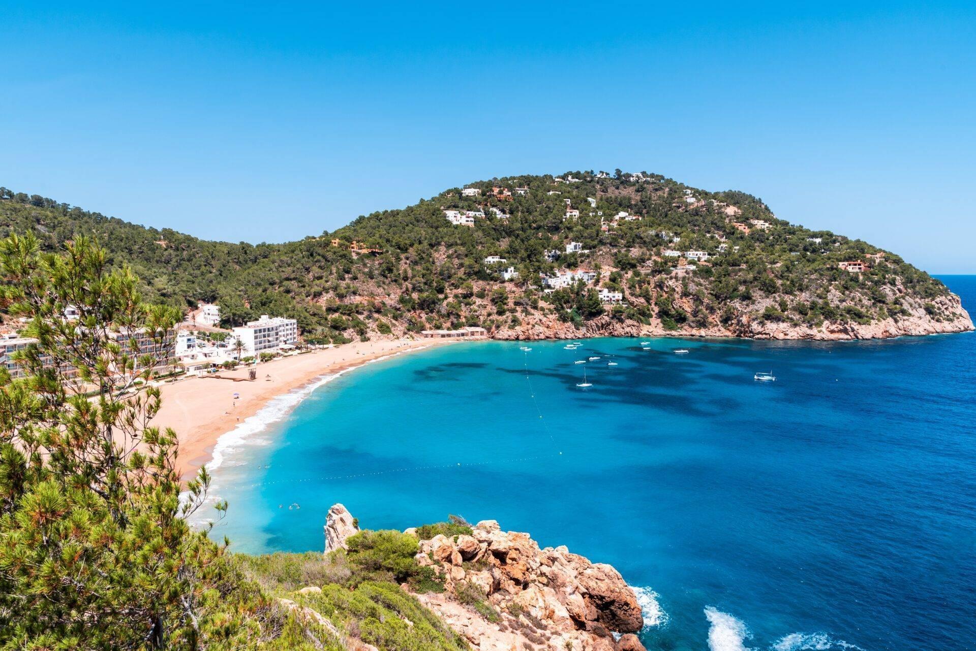 Une baie méditerranéenne aux eaux turquoise, bordée d’une plage de sable fin et de falaises rocheuses boisées avec habitations