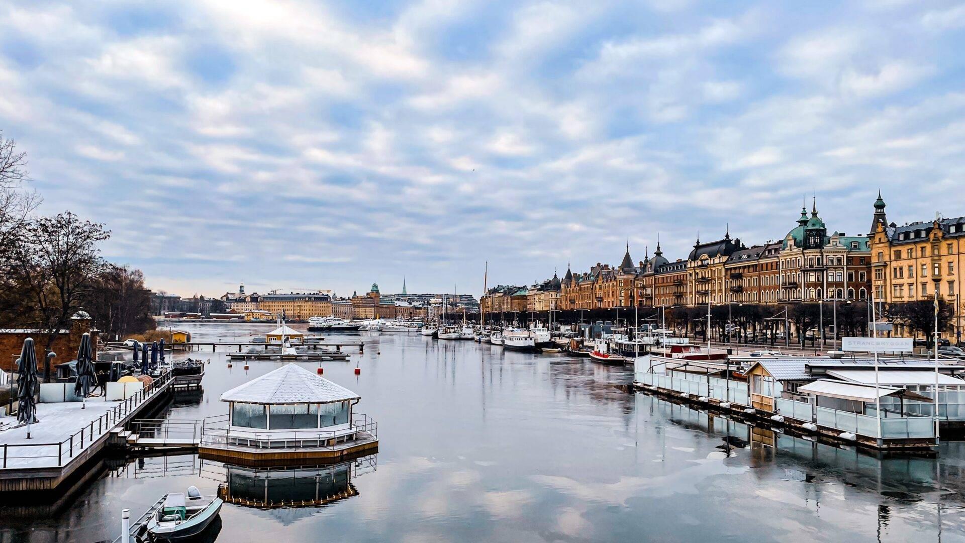 Vue hivernale d’un port scandinave avec bateaux amarrés, architecture colorée et eau gelée reflétant le ciel nuageux