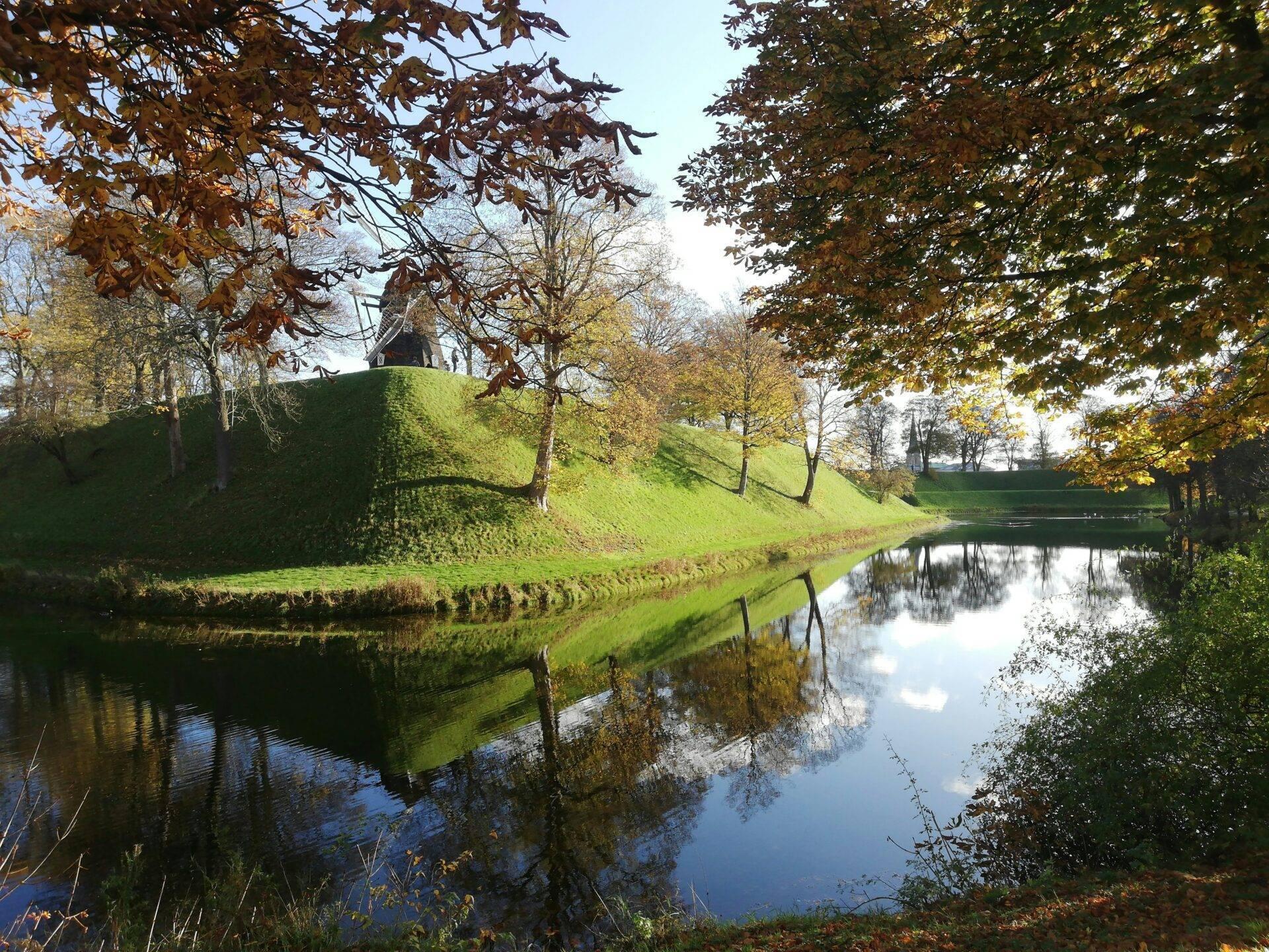 Un château fort sur une butte verdoyante entouré d’un fossé d’eau reflétant les arbres d’automne