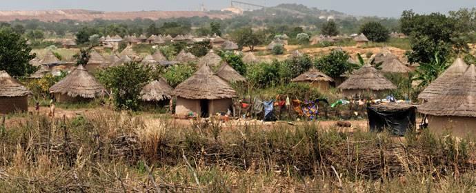 Village traditionnel avec cases aux toits de chaume, champs cultivés et habitants dans une région rurale d’Afrique de l’Ouest