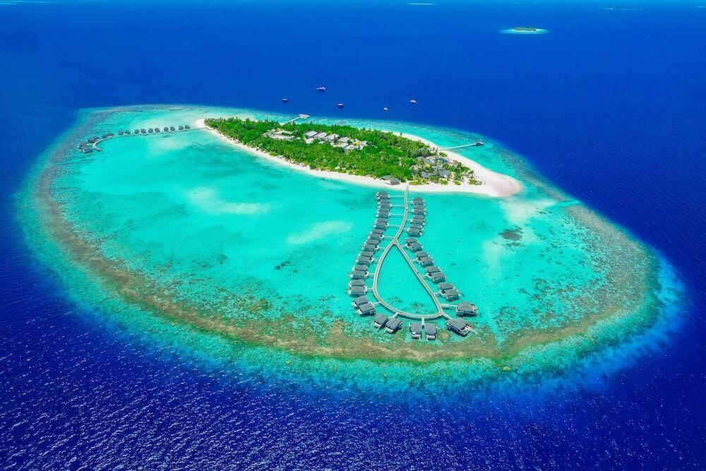 Vue aérienne d’un atoll tropical avec île verdoyante, bungalows sur pilotis et lagon turquoise cristallin