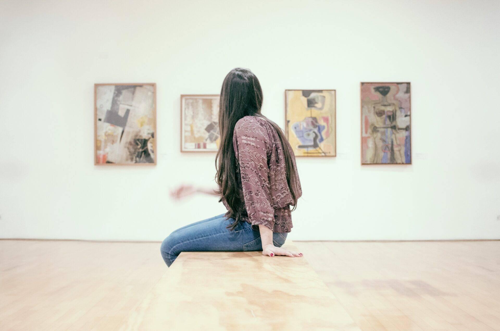 Une femme assise contemple des tableaux abstraits dans une galerie d’art moderne aux murs blancs