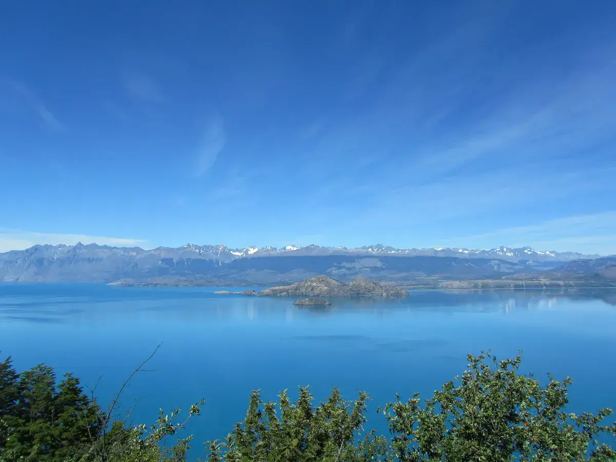 Lac alpin turquoise avec îlots rocheux et chaîne montagneuse enneigée en arrière-plan sous ciel bleu