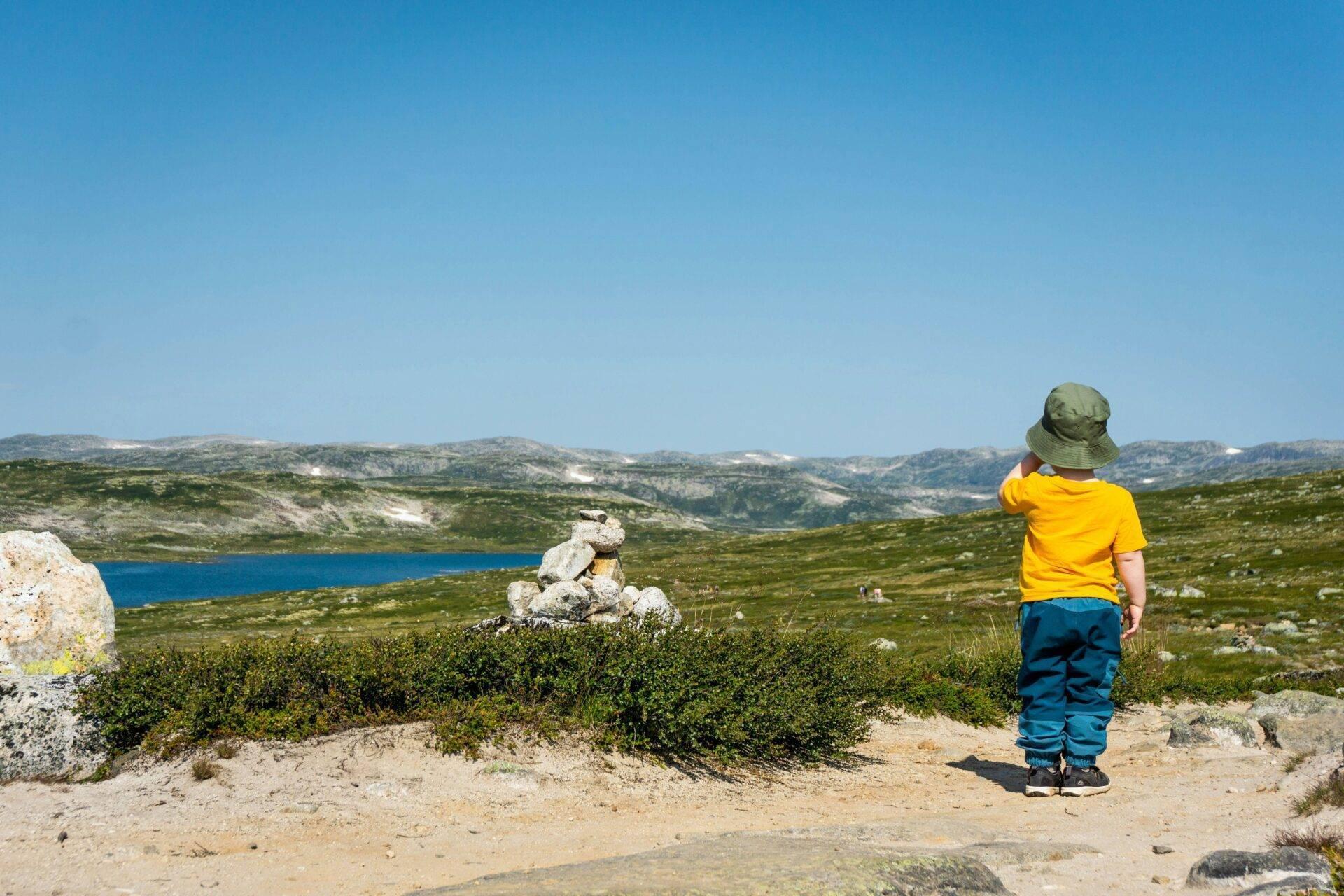 Un enfant en vêtements colorés observe un paysage montagneux alpin avec lac bleu et cairn de pierres