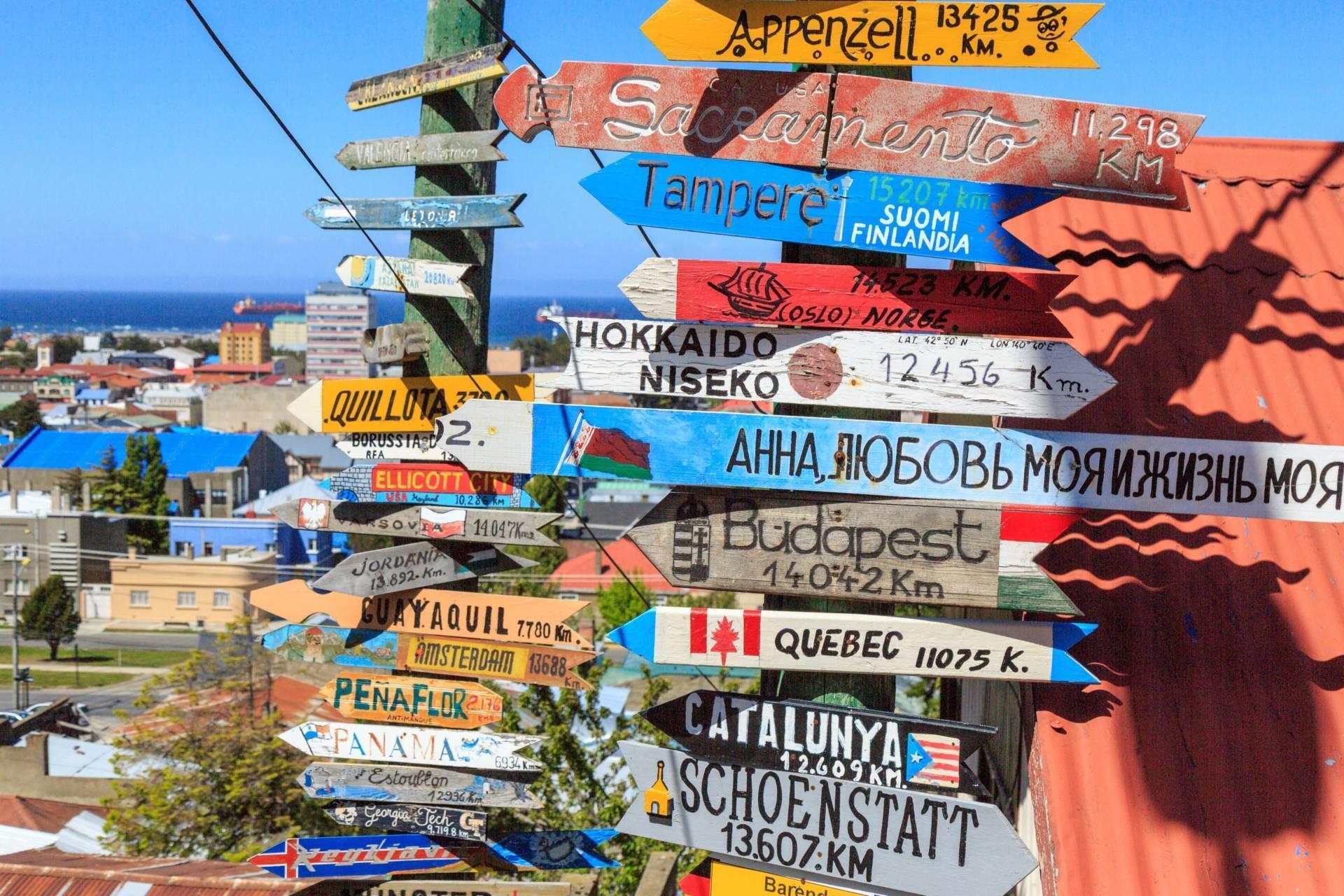 Un poteau directionnel coloré avec panneaux indiquant des destinations du monde entier, avec vue côtière et bâtiments en arrière-plan