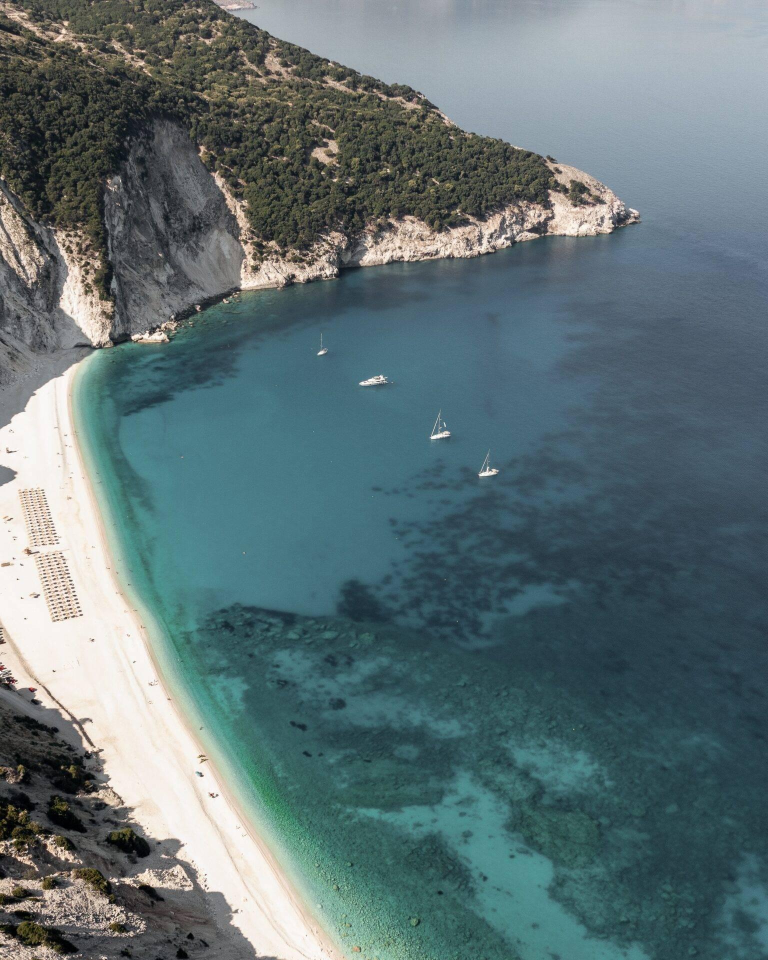 Vue aérienne d’une plage de sable blanc bordée de falaises calcaires avec eau turquoise et voiliers ancrés