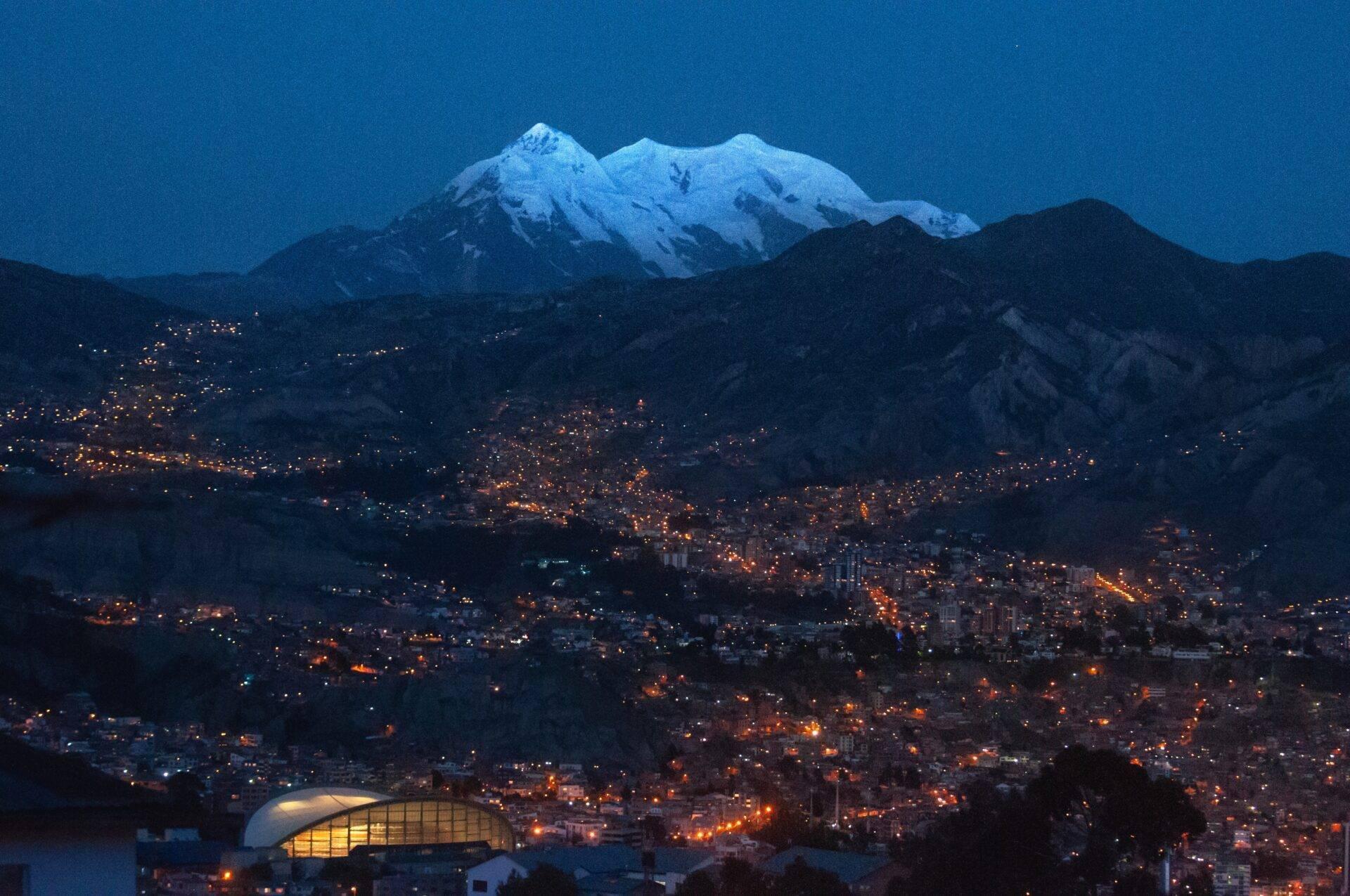 Ville illuminée de nuit au pied d’un volcan enneigé sous ciel bleu crépusculaire, avec stade moderne en premier plan