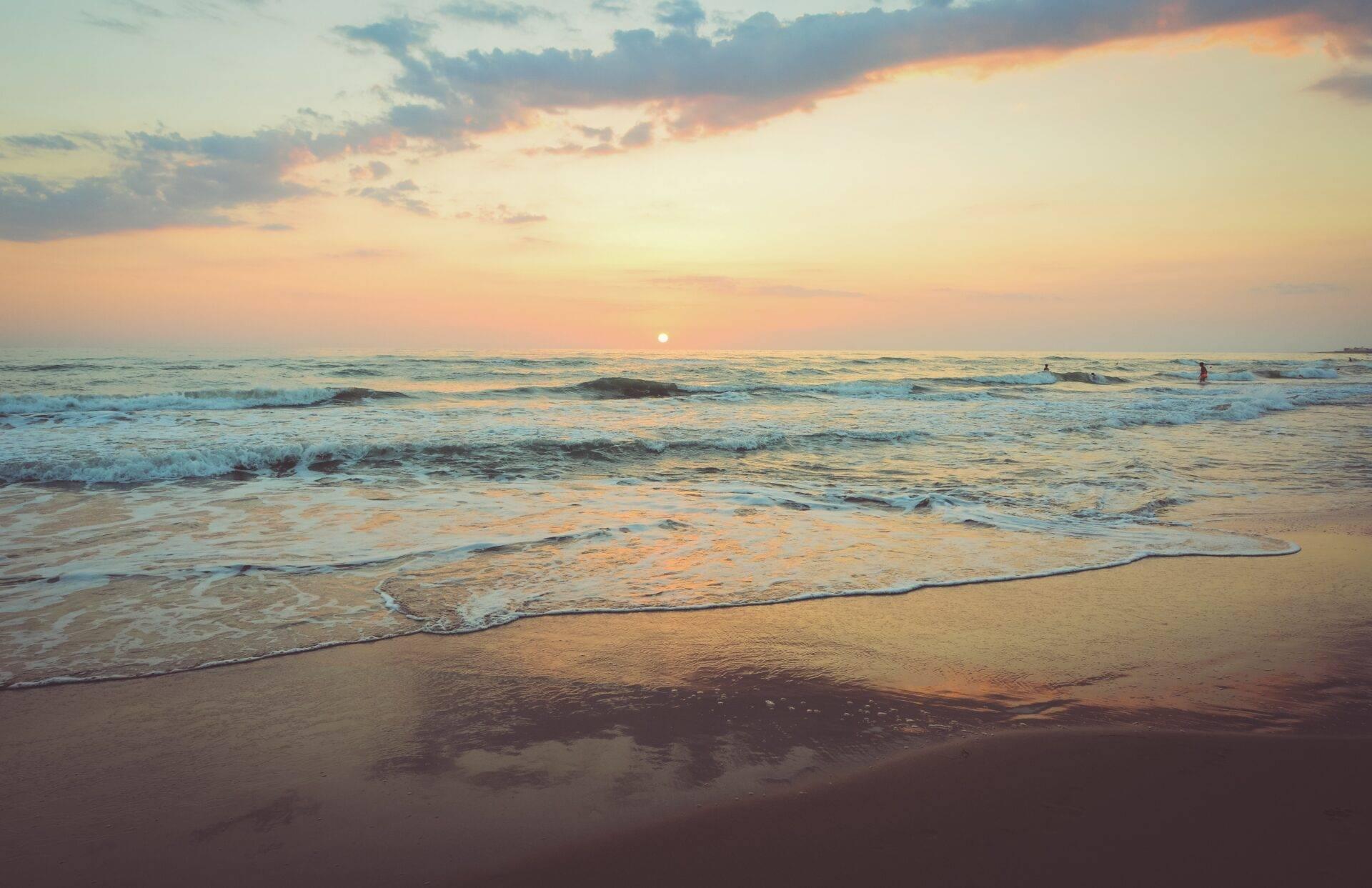 Coucher de soleil sur une plage de sable avec vagues et quelques baigneurs à l’horizon