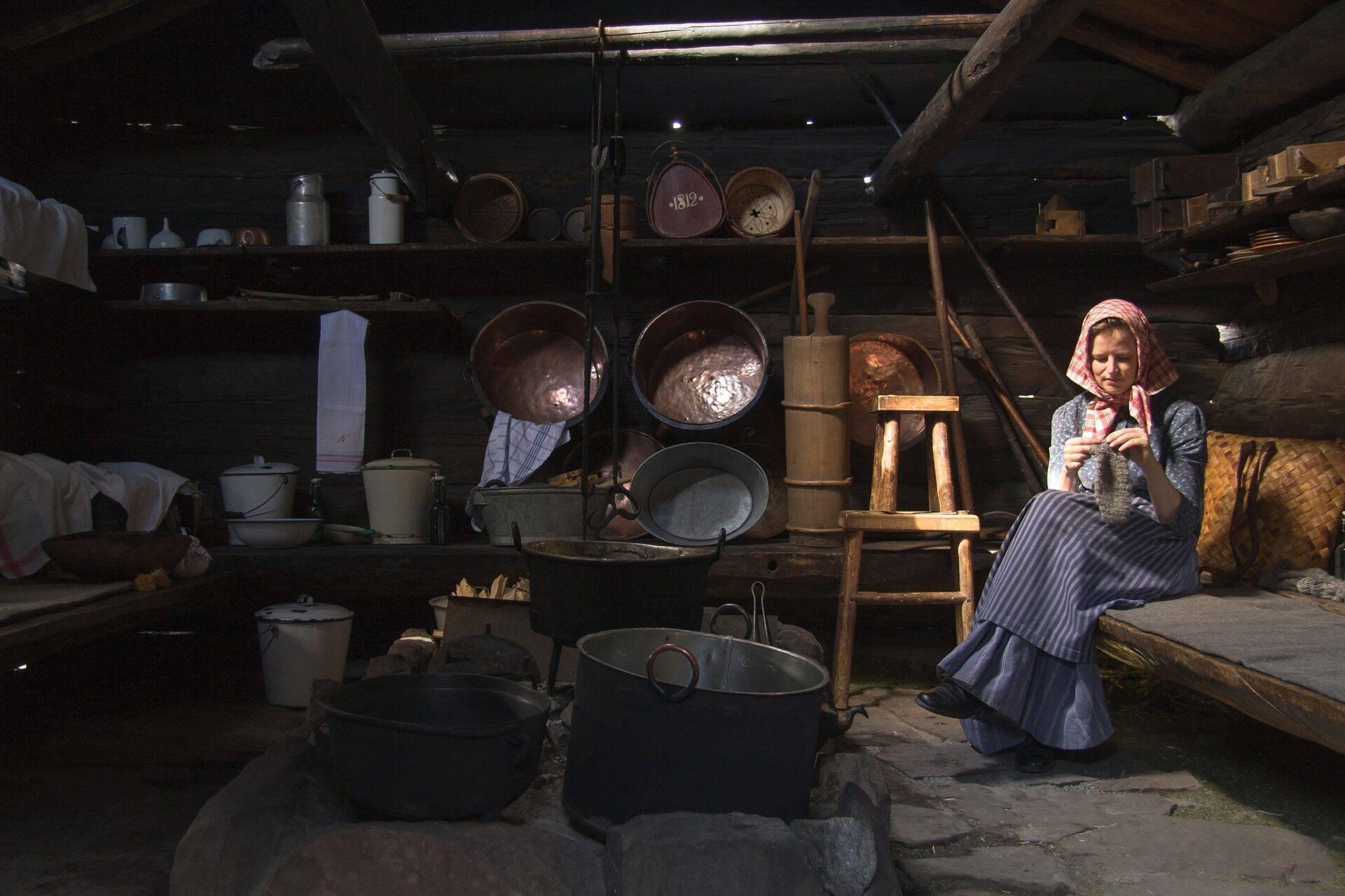 Une femme en costume traditionnel travaille dans une cuisine historique avec ustensiles et mobilier d’époque