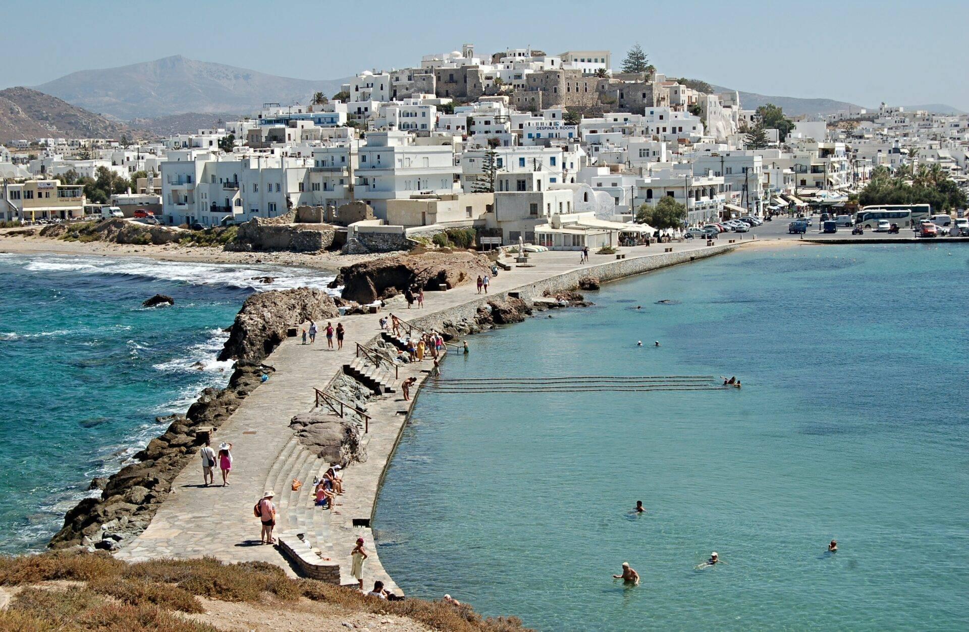 Un village côtier méditerranéen blanc perché sur une colline avec plage turquoise et promenade rocheuse animée