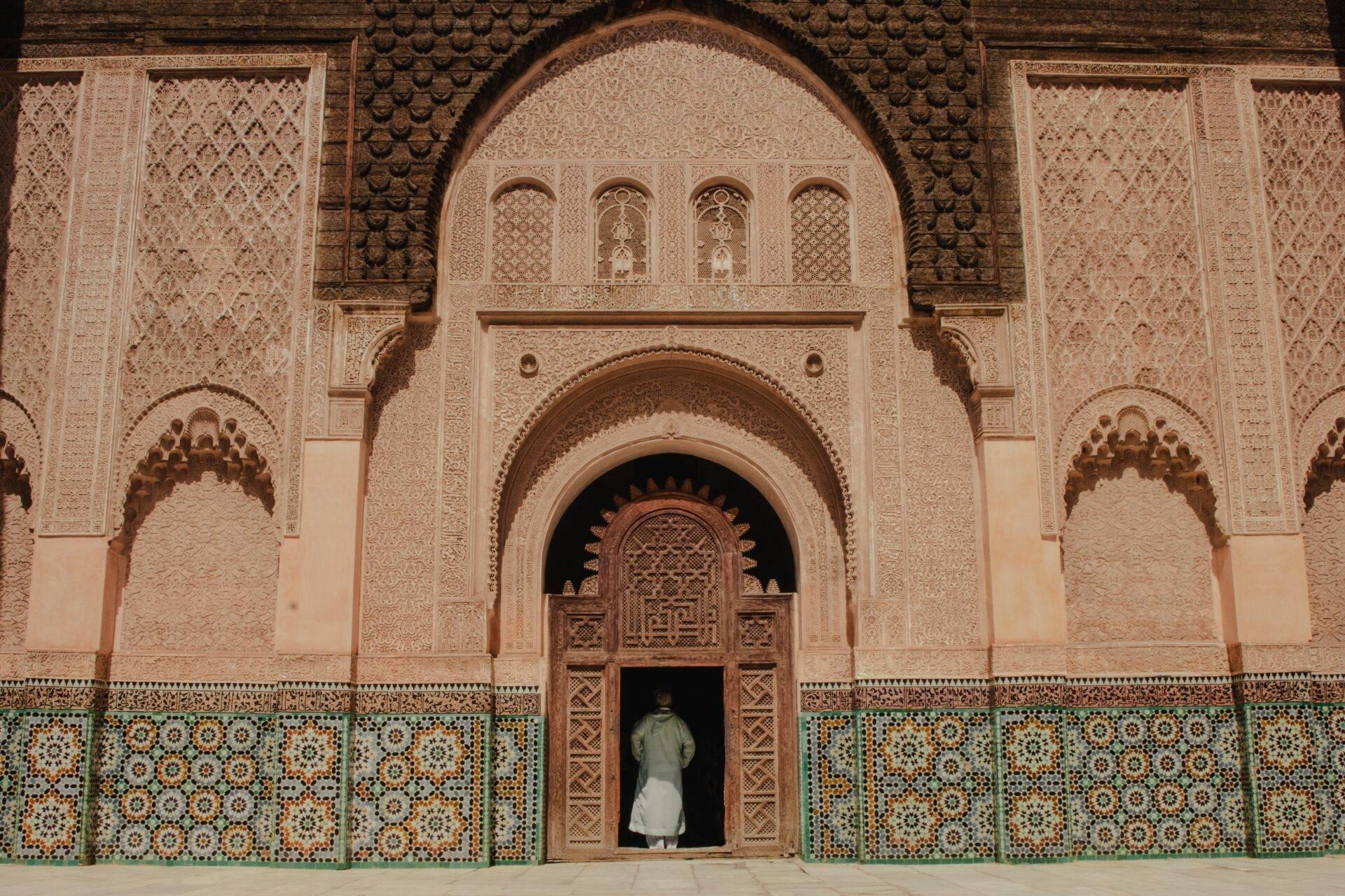 Une personne en vêtements blancs devant l’entrée ornée d’une mosquée marocaine aux détails géométriques et carrelages turquoise