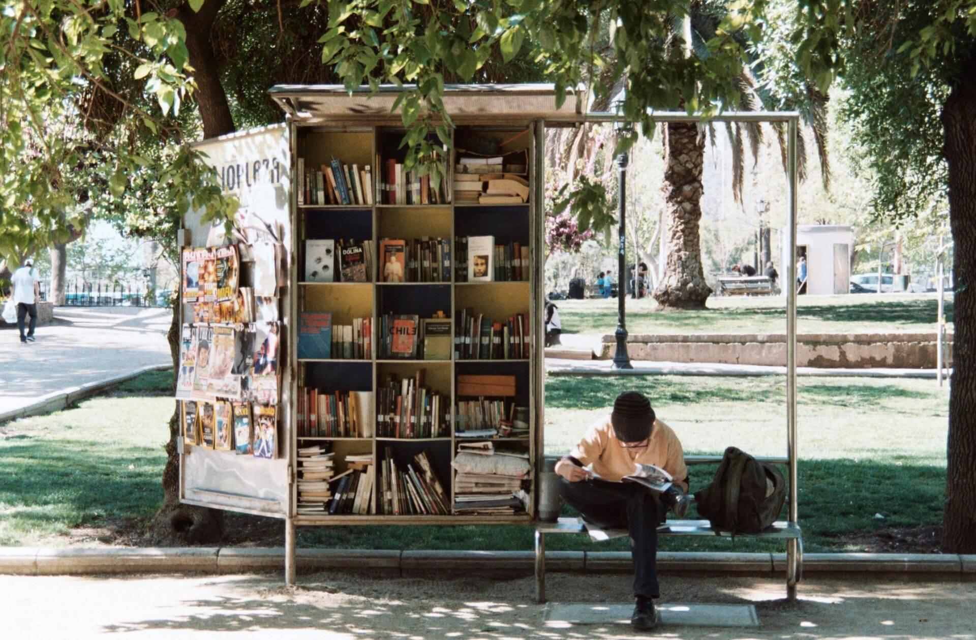 Un homme lit assis devant une petite bibliothèque publique en plein air sous des arbres dans un parc urbain
