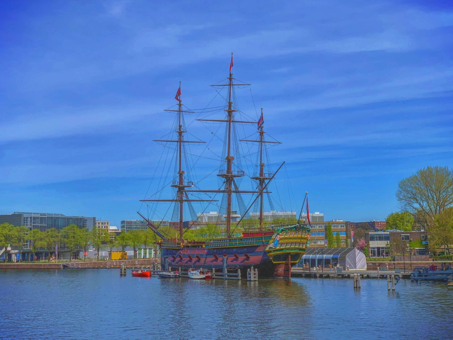 Un navire trois-mâts coloré amarré dans un port fluvial urbain sous un ciel bleu dégagé
