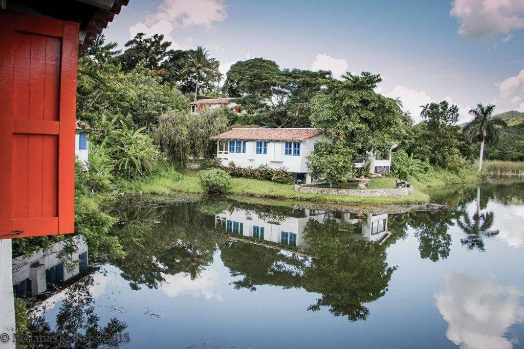 Maisons coloniales blanches et bleues avec porte rouge se reflétant dans un canal tropical entouré de végétation luxuriante