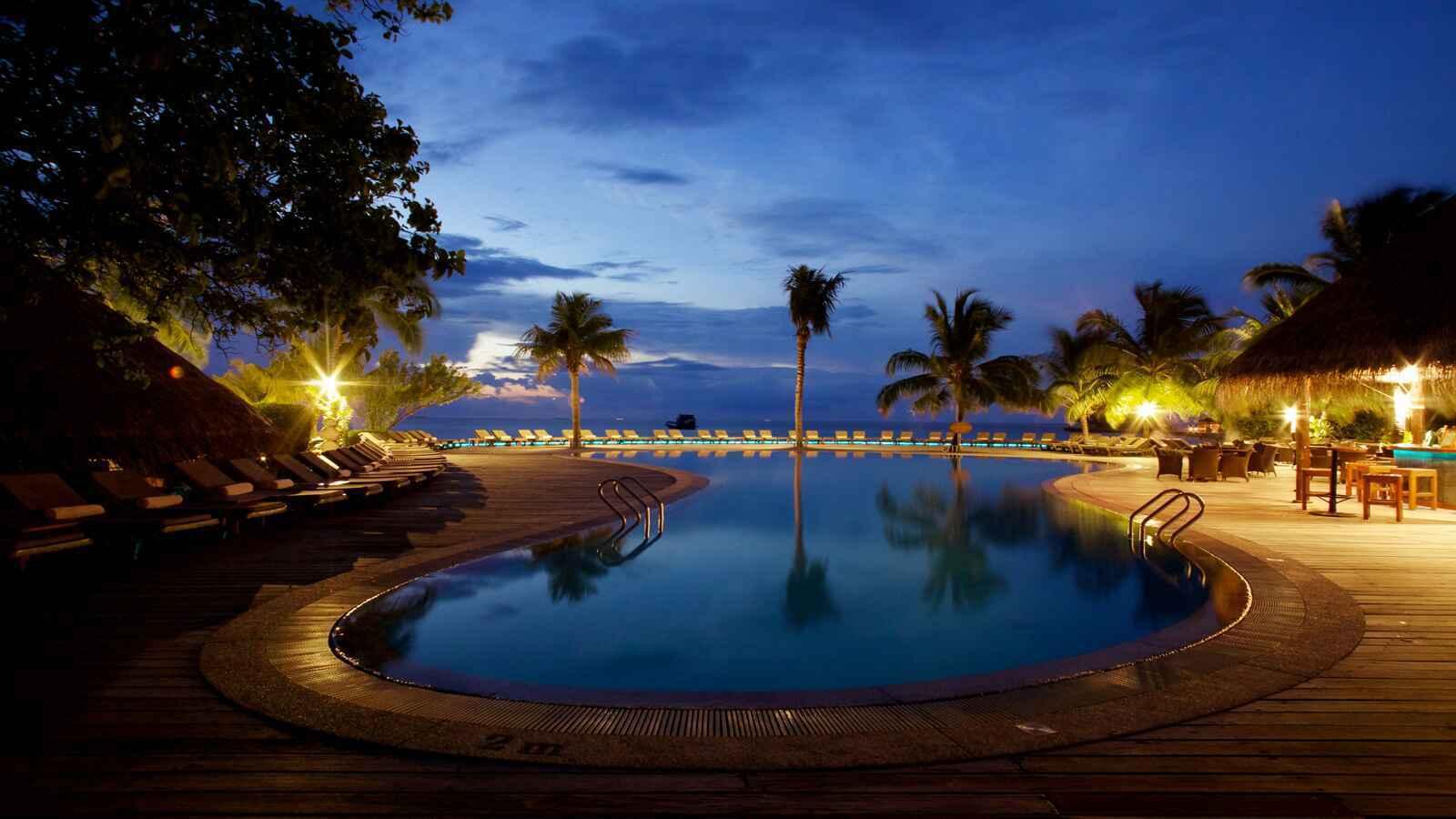 Une piscine infinie luxueuse illuminée au crépuscule avec palmiers et transats face à l’océan