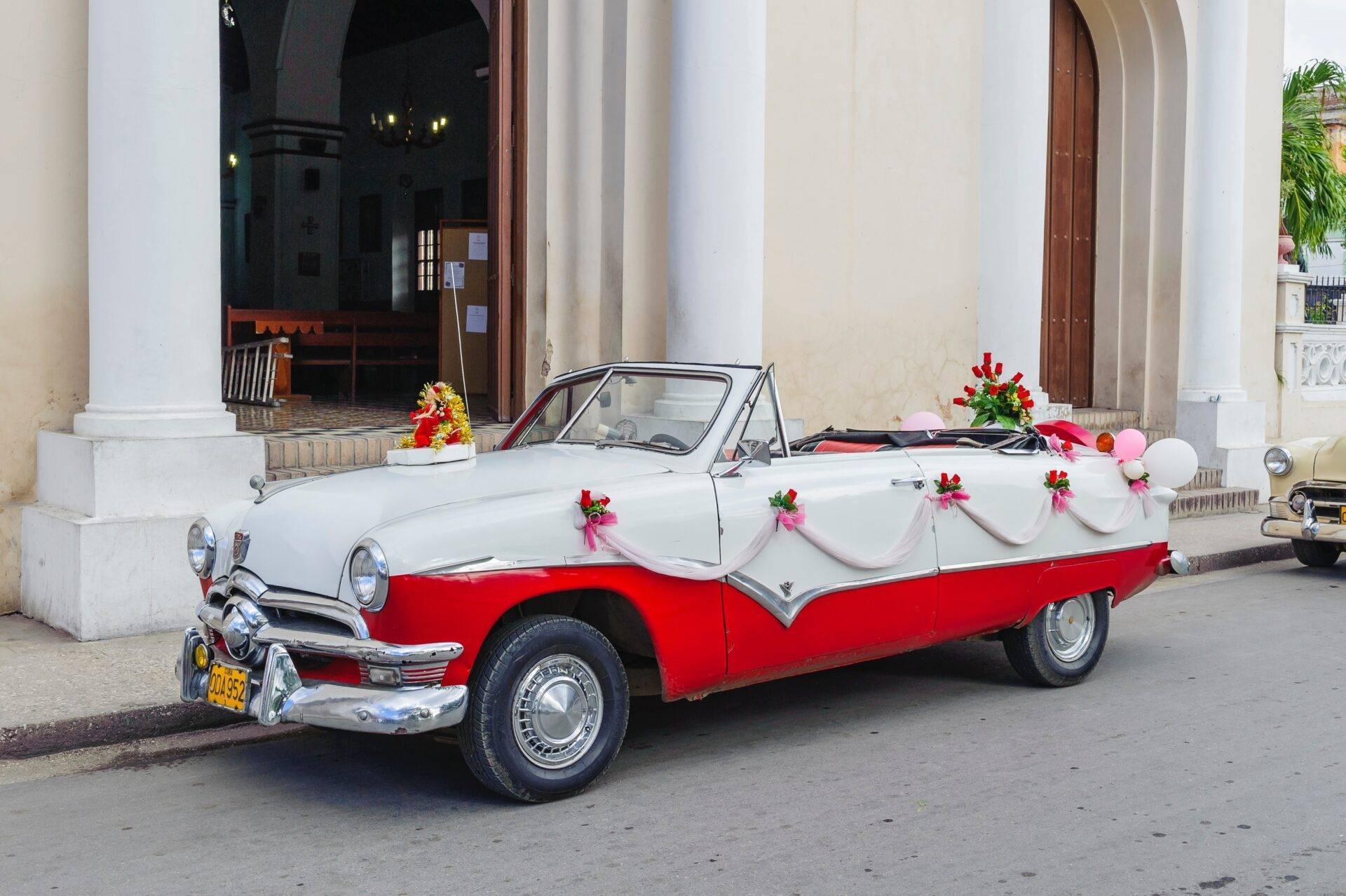 Une voiture décapotable rouge et blanche décorée de fleurs roses stationnée devant un bâtiment colonial blanc