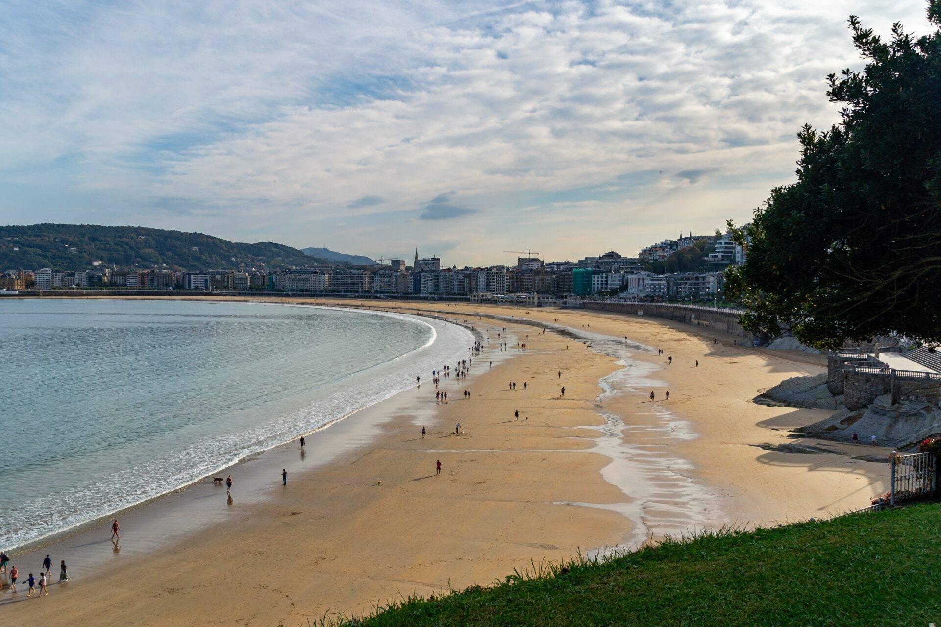 Vue panoramique d’une grande plage de sable fin avec baigneurs et une ville côtière en arrière-plan sous ciel nuageux