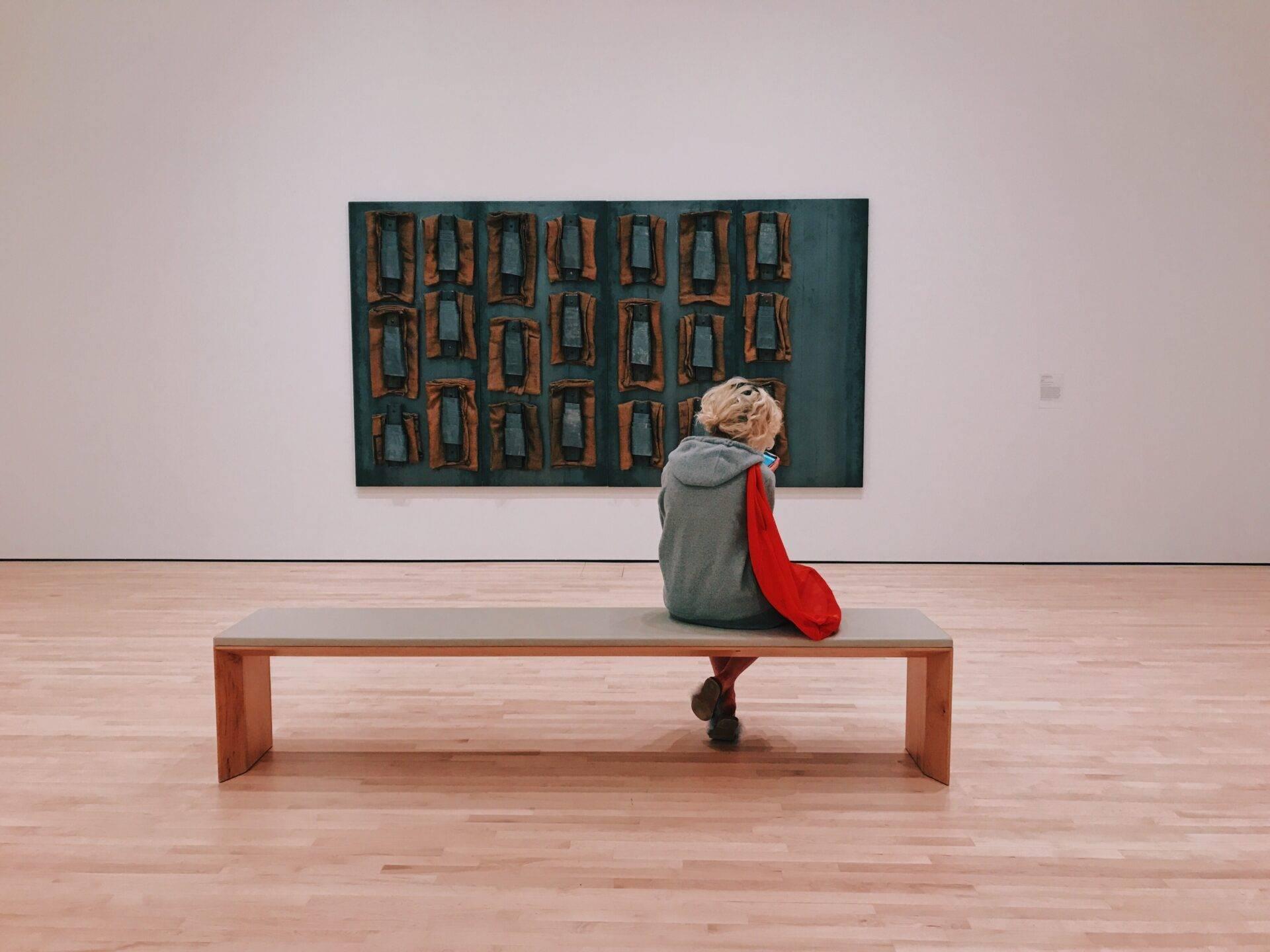 Une personne assise sur un banc en bois contemple une peinture abstraite aux tons bleu et orange dans une galerie d’art