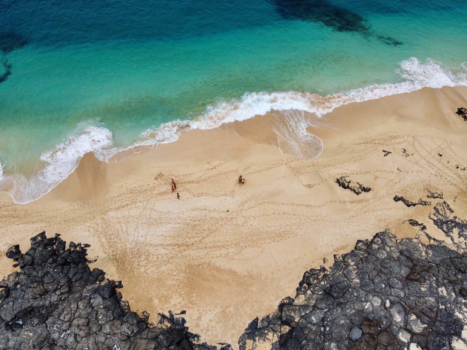 Vue aérienne d’une plage de sable fin avec vagues turquoise, rochers noirs encadrant le rivage désert