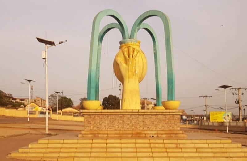 Monument public avec arches vertes et main jaune centrale dans une place urbaine aride