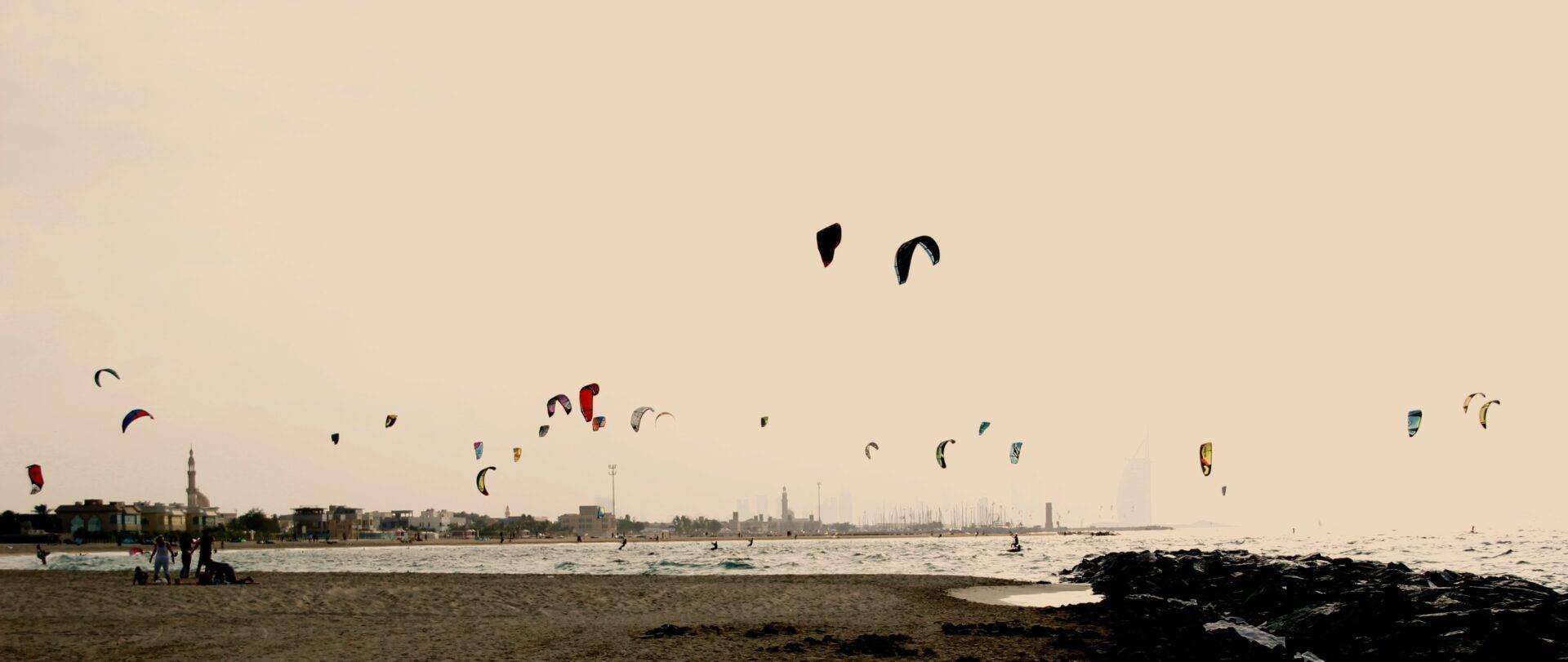 Une plage avec de nombreux kitesurfeurs dans l’eau et des voiliers amarrés, face à une ville côtière