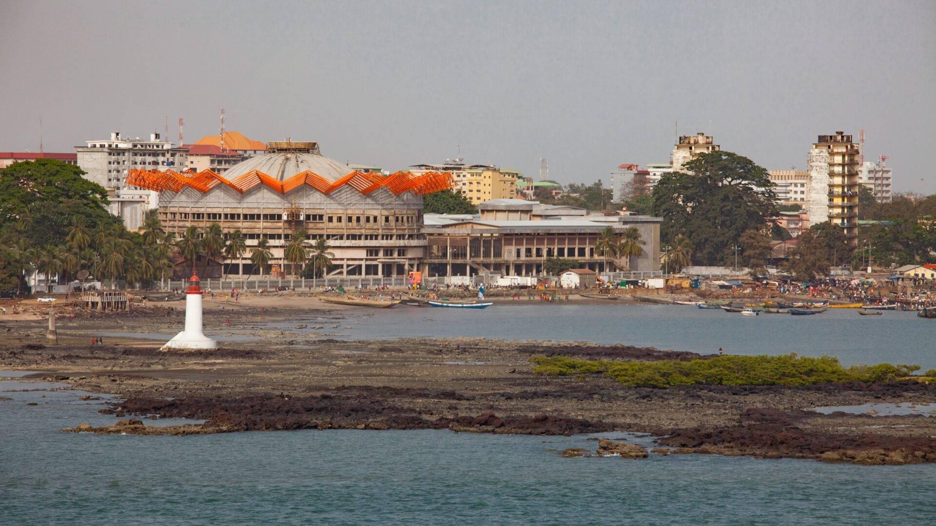 Vue côtière d’une ville africaine avec un bâtiment aux toits orange ondulés, un phare blanc et des bateaux de pêche