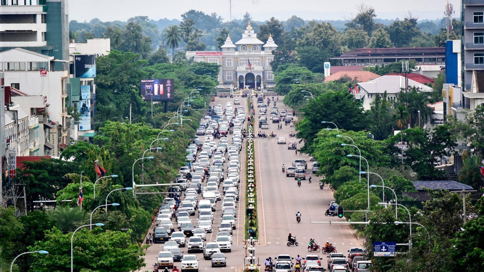 Vue aérienne d’une large avenue bordée de voitures avec un temple bouddhiste blanc au fond, entouré de végétation luxuriante