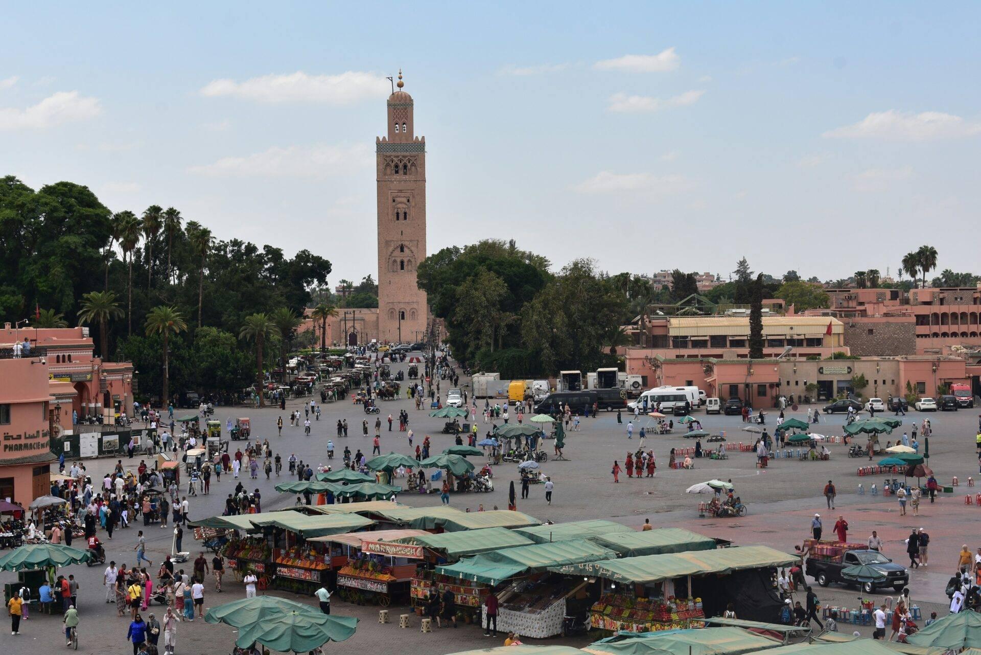 Une grande place animée avec mosquée historique, marchands sous des auvents verts, foule de visiteurs et bâtiments ocre traditionnels