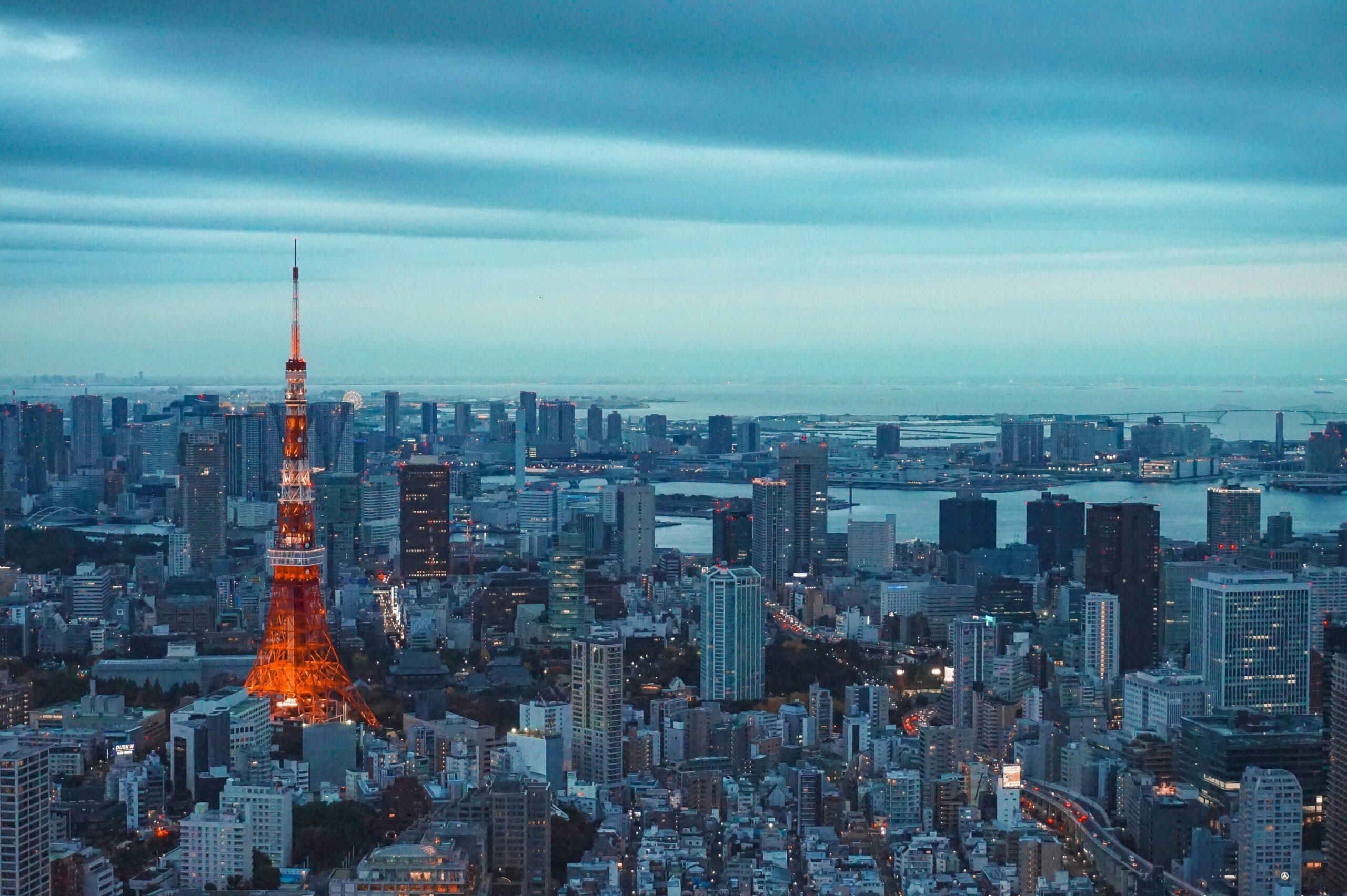 Vue aérienne d’une métropole japonaise au crépuscule avec une tour illuminée en rouge dominant les gratte-ciel et la baie