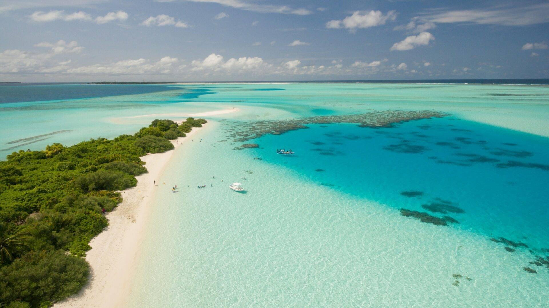 Vue aérienne d’une île tropicale avec plage de sable blanc, lagon turquoise cristallin et végétation luxuriante, quelques bateaux visibles