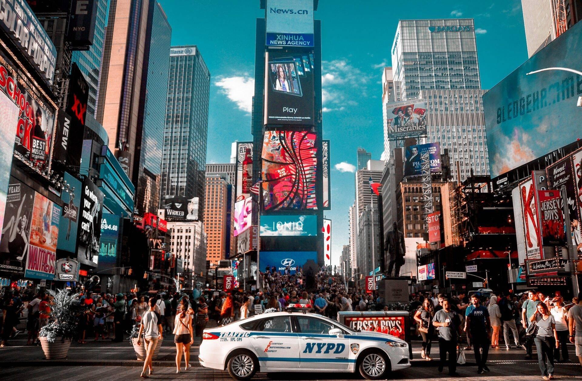 Une rue animée avec des gratte-ciel, des panneaux publicitaires géants et une voiture de police, sous un ciel bleu dégagé