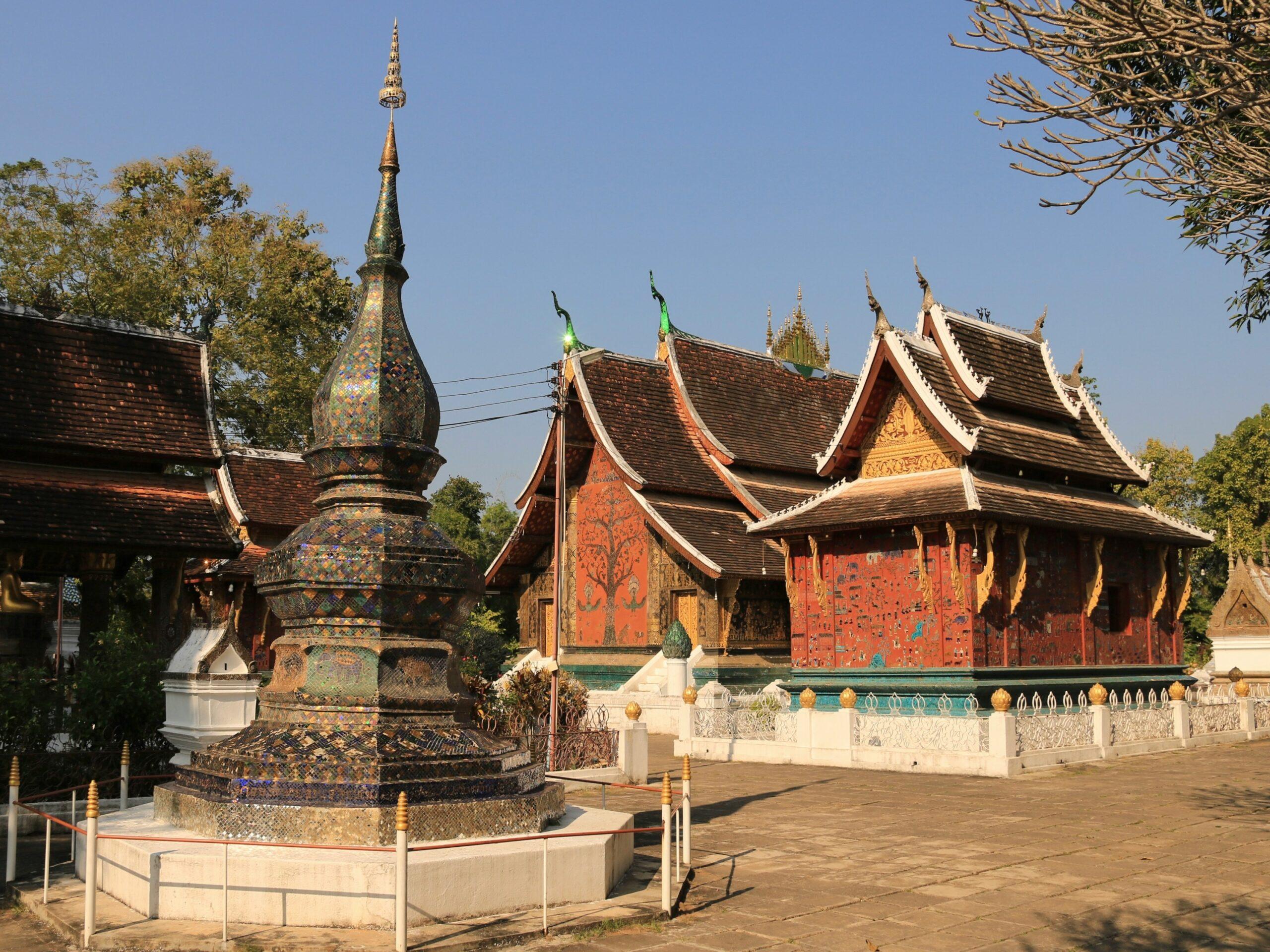 Un temple bouddhiste traditionnel avec toits recourbés dorés, stupa orné et bâtiments rouges, entouré d’arbres